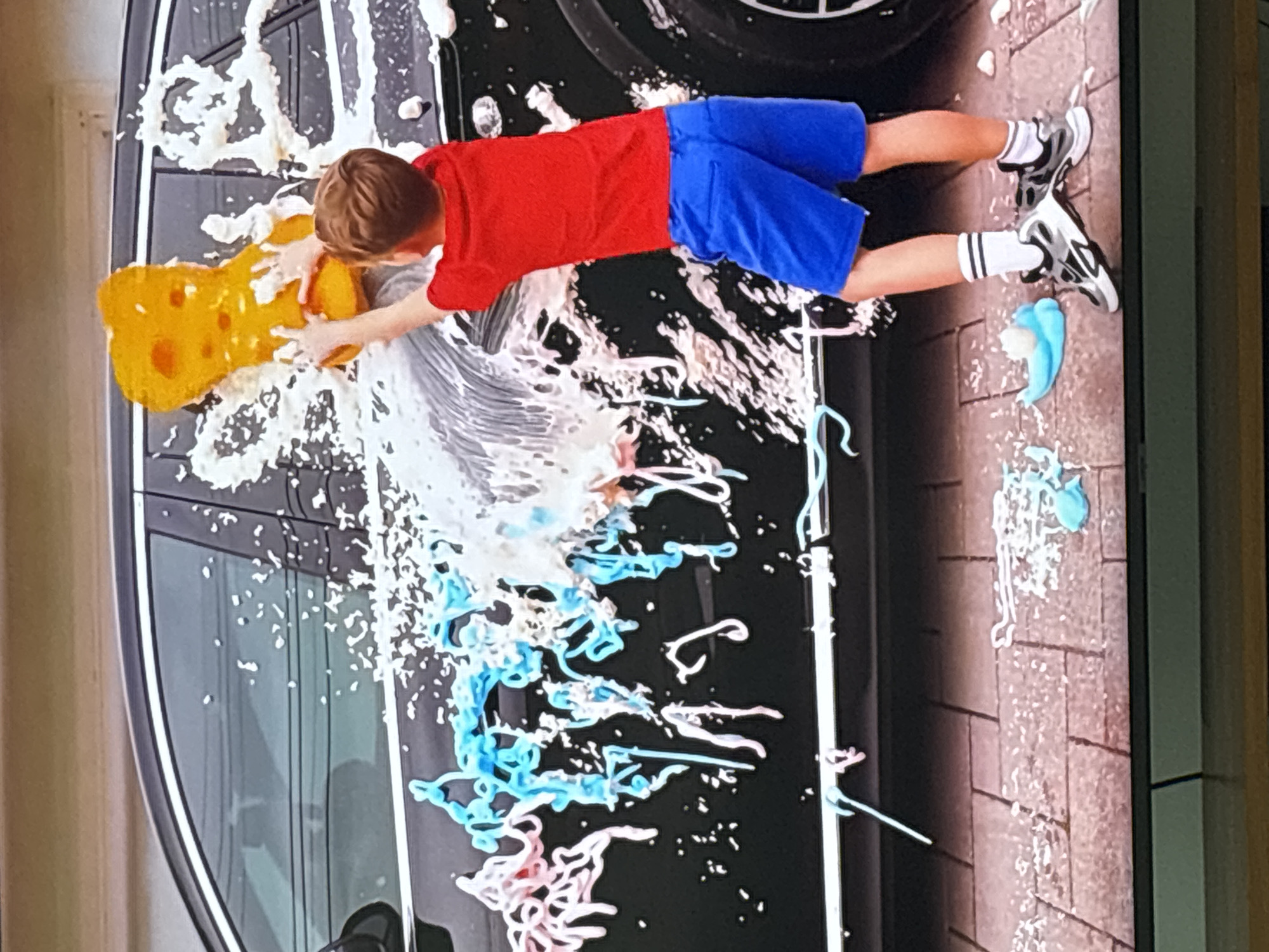 A young boy in a red t-shirt and blue shorts is washing a black car. He is holding a yellow sponge in his hands and applying it to the car door, creating a lot of white foam. The car is covered in colorful soap suds, some of which are in swirls and strands. The boy is standing on a paved driveway with a brick pattern. There are also some blue and white toy-like objects on the ground. The background shows a garage door or wall. The image is a medium shot from the side, focusing on the boy and the car. The location is not specified.