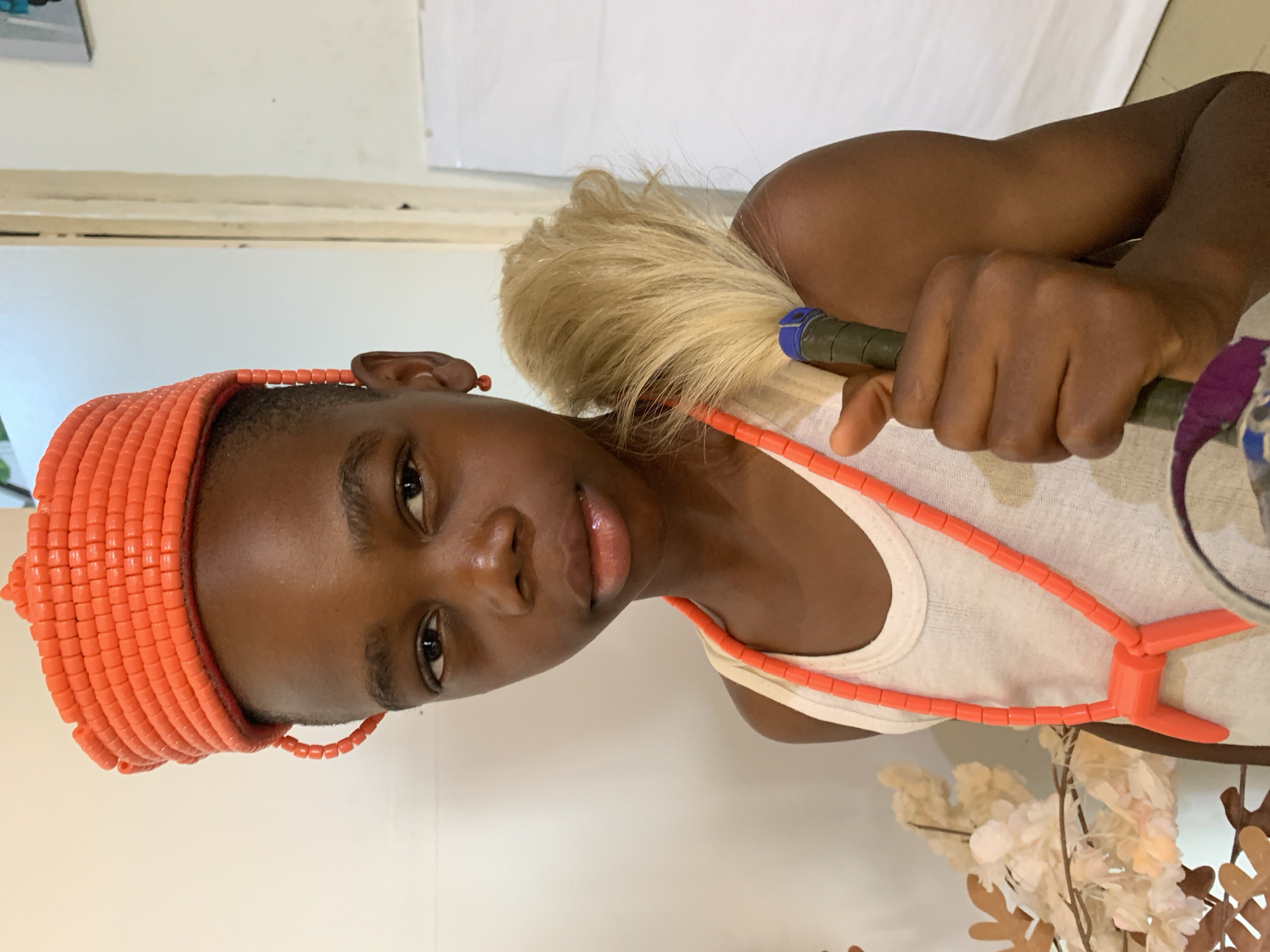 A young person is shown from the chest up, wearing a traditional Nigerian outfit. They have on a vibrant orange beaded crown and a matching beaded necklace. A single orange bead earring is visible in their left ear. They are also wearing a simple white tank top. In their right hand, they are holding a whisk with a blonde tassle and a dark grip. The background is a plain wall, suggesting an indoor setting. The lighting is soft and diffused, creating a warm glow. The image does not provide enough information to determine the specific location in Jos, Nigeria.