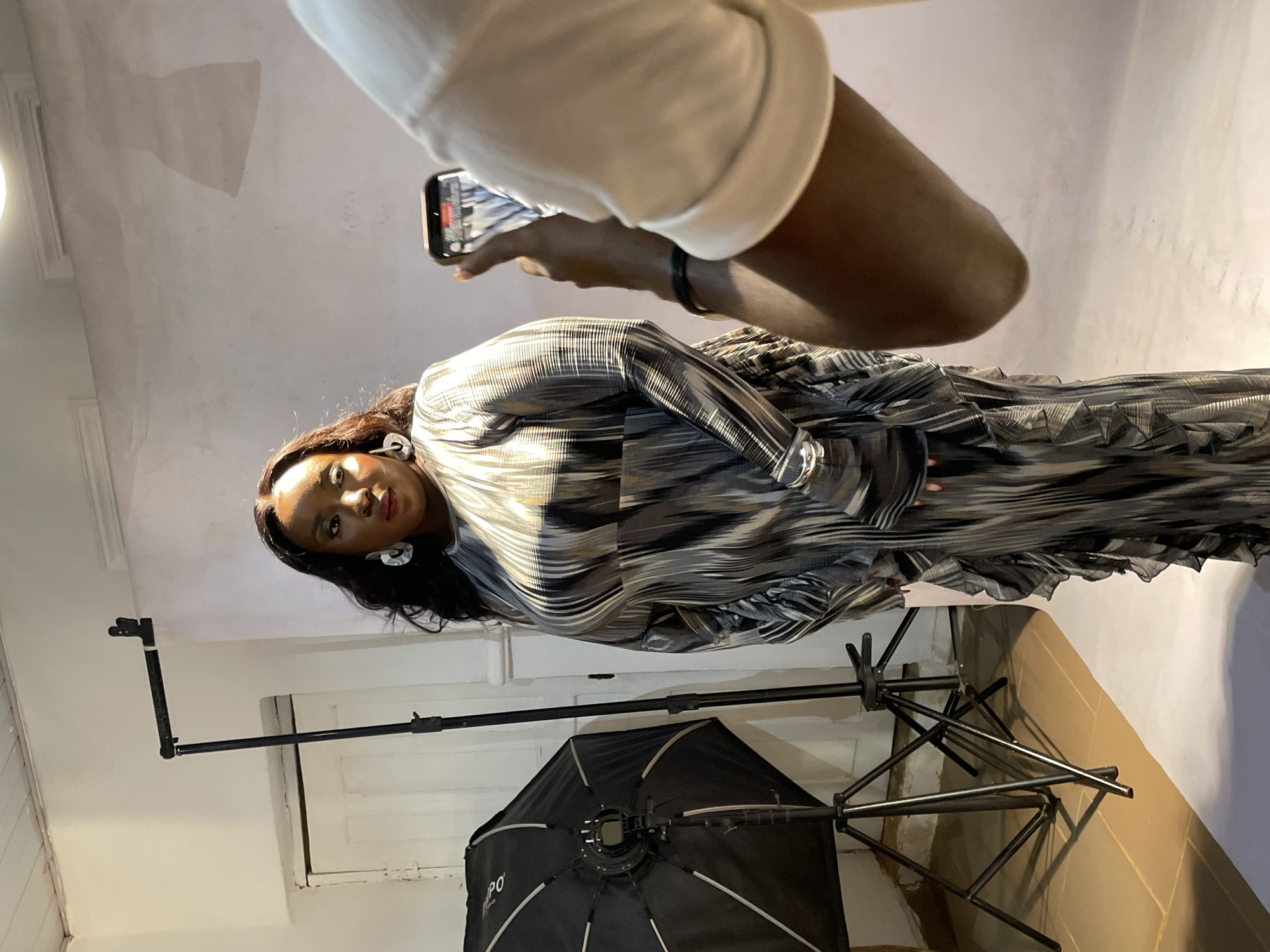 This image captures an indoor photoshoot or video recording session taking place in Jos, Nigeria. The scene is oriented vertically, showing a low-angle perspective looking upwards.
The main subject is a woman with dark hair, styled down, wearing a striking long-sleeved dress. The dress features a complex pattern of horizontal stripes in black, white, and various shades of grey, with a pleated or ruffled texture, especially noticeable on the skirt and sleeves. She is also adorned with prominent, white, circular or geometric earrings. Her gaze is directed towards the camera, maintaining a neutral to slightly serious expression.
In the foreground, partially visible, is a person holding a smartphone horizontally, seemingly capturing the ongoing session. Only the person's right arm, wearing a white long-sleeved top, and the back of the smartphone are clearly discernible. The phone's screen displays what appears to be a live feed or a recent capture, mirroring the patterned dress of the subject.
The setting is an indoor space, likely a studio or a versatile room, characterized by light-colored walls that exhibit some textural imperfections or wear. The floor is covered with light brown or beige tiles. Essential photography equipment is present, including a large black softbox or light modifier positioned on a stand to the left of the woman, indicating a controlled lighting environment. The base of another stand or tripod is visible near the floor. The letters "PO" are visible on the softbox, suggesting a brand name. The overall atmosphere suggests a professional creative activity.