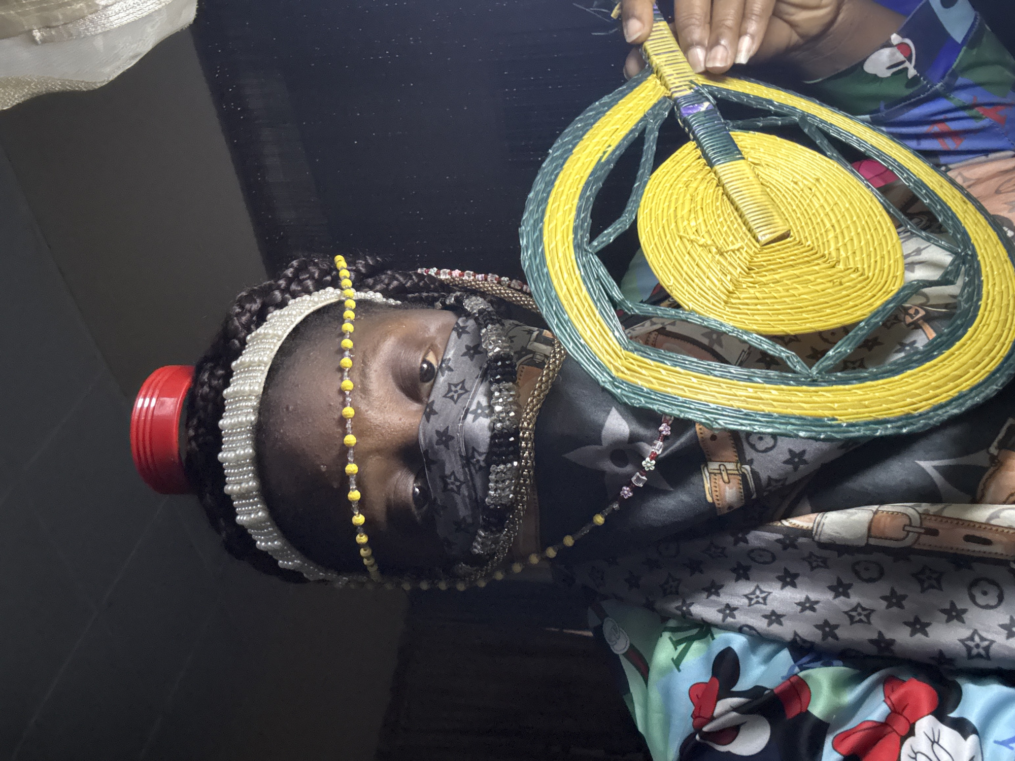 A person, seemingly female, is holding a round, woven fan. She is wearing beaded necklaces and a pearl headband, as well as a patterned face mask. She is surrounded by a collection of patterned fabrics, including those with cartoon characters and designer logos. The image's location is not discernible from the visual data provided. No location of Madangala, Nigeria is evident.