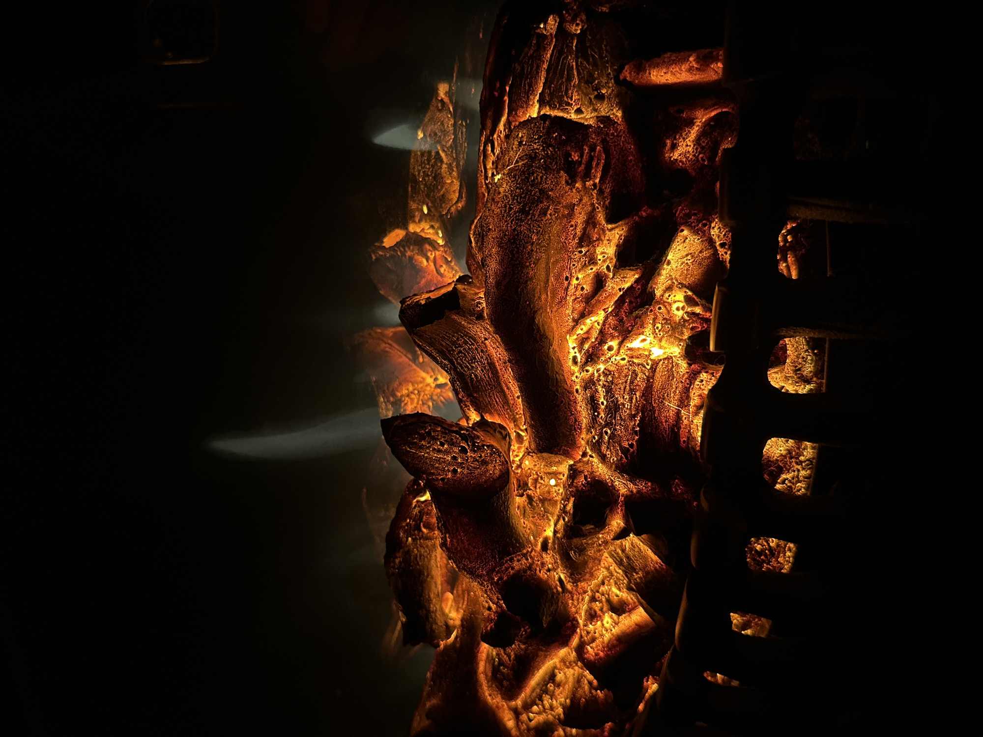 This is a close-up, low-angle, full-shot photograph of glowing embers and artificial logs in a fireplace. The logs are dark brown with visible textures and are illuminated by a bright orange and yellow glow, suggesting heat. Some wisps of white smoke or light are visible rising from the logs. In the foreground, a dark metal grate with arched openings partially obscures the view of the embers. The background is entirely black, creating a stark contrast with the illuminated logs. The scene appears to be indoors, likely in a home or commercial setting. There are no people or animals present.