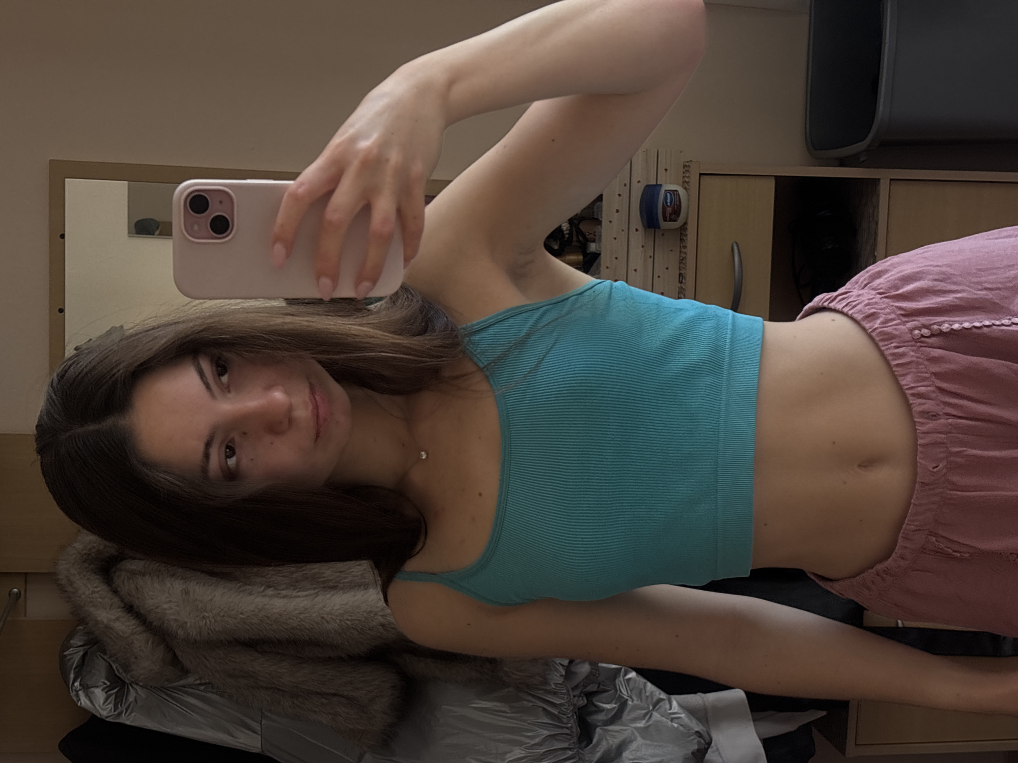 The media file is a selfie taken by a young woman indoors, likely in a bedroom or dressing area. She is holding up a smartphone to take the picture, which is framed to show her upper body and a portion of her face. She has long, dark brown hair and is wearing a teal, ribbed tank top and pink, ruffled shorts. A small diamond necklace is visible around her neck.
In the background, a mirror reflects part of the room, including what appears to be a wooden dresser or cabinet. There are also some personal items visible, such as a jar of Vaseline and clothing draped over furniture, including a fuzzy grey coat and a silver jacket. The lighting suggests it is daytime, with artificial indoor lighting. The woman's expression is neutral, and the overall mood is casual and relaxed. There is no visible text in the image. The location is identified as Varna, Bulgaria, but this context is not visually evident within the image itself.