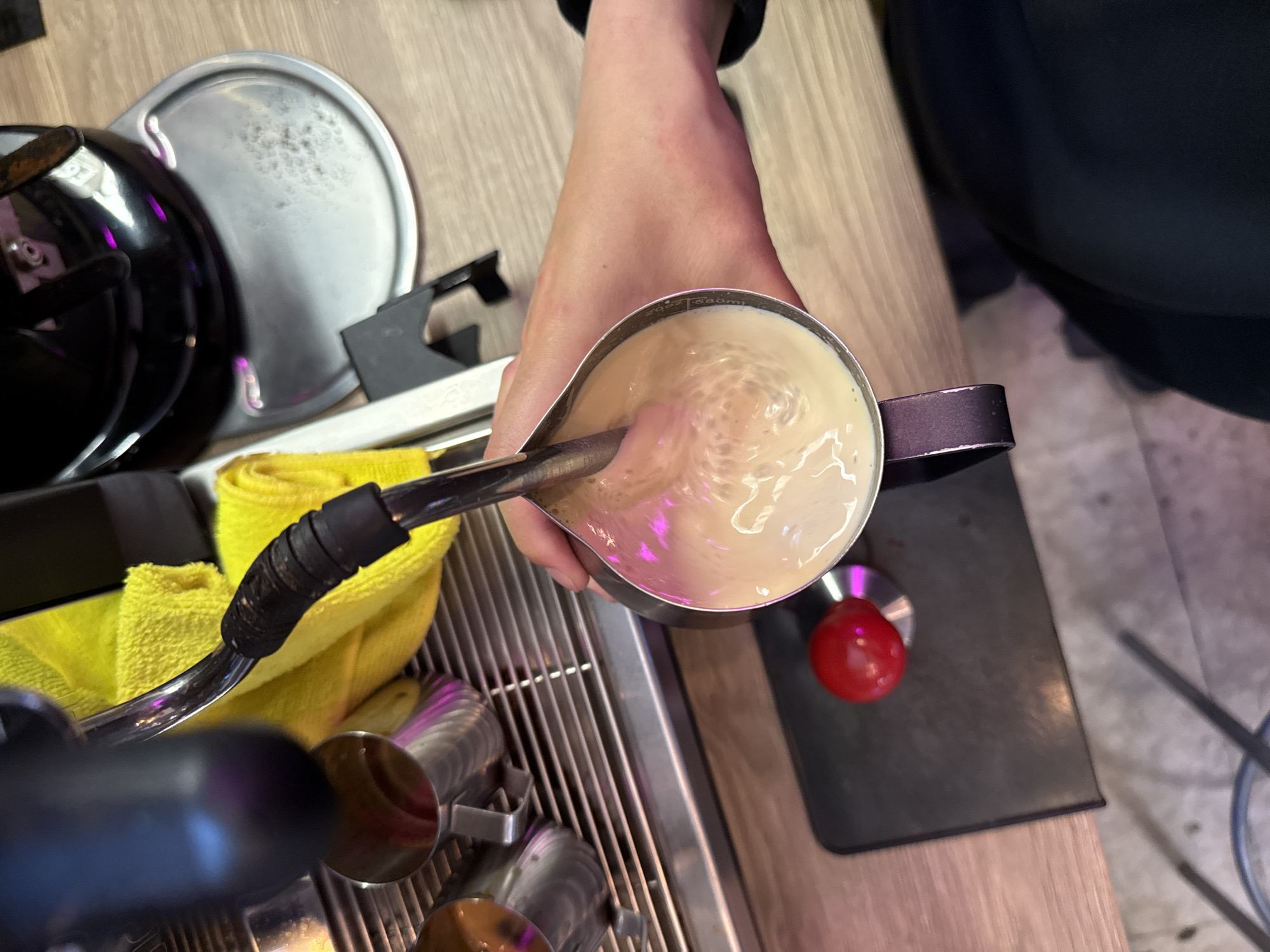 A close-up overhead shot shows a person's hand holding a metal pitcher filled with frothy, light-brown liquid, likely milk being steamed for coffee. The pitcher is tilted, and the liquid is swirling, with bubbles visible on the surface. A steaming wand from an espresso machine is partially submerged in the liquid, indicating the milk is being heated and aerated. In the background, a yellow microfiber cloth is draped over part of the espresso machine. To the left, several stainless steel cups are visible on the machine's drip tray. Below the pitcher, a black mat with a red, round object, possibly a tamper, is placed on the counter. The counter surface appears to be a light-colored wood grain. The lighting suggests an indoor setting, possibly a cafe or a home kitchen. There are no specific indicators to place this scene in Moscow, Russia.