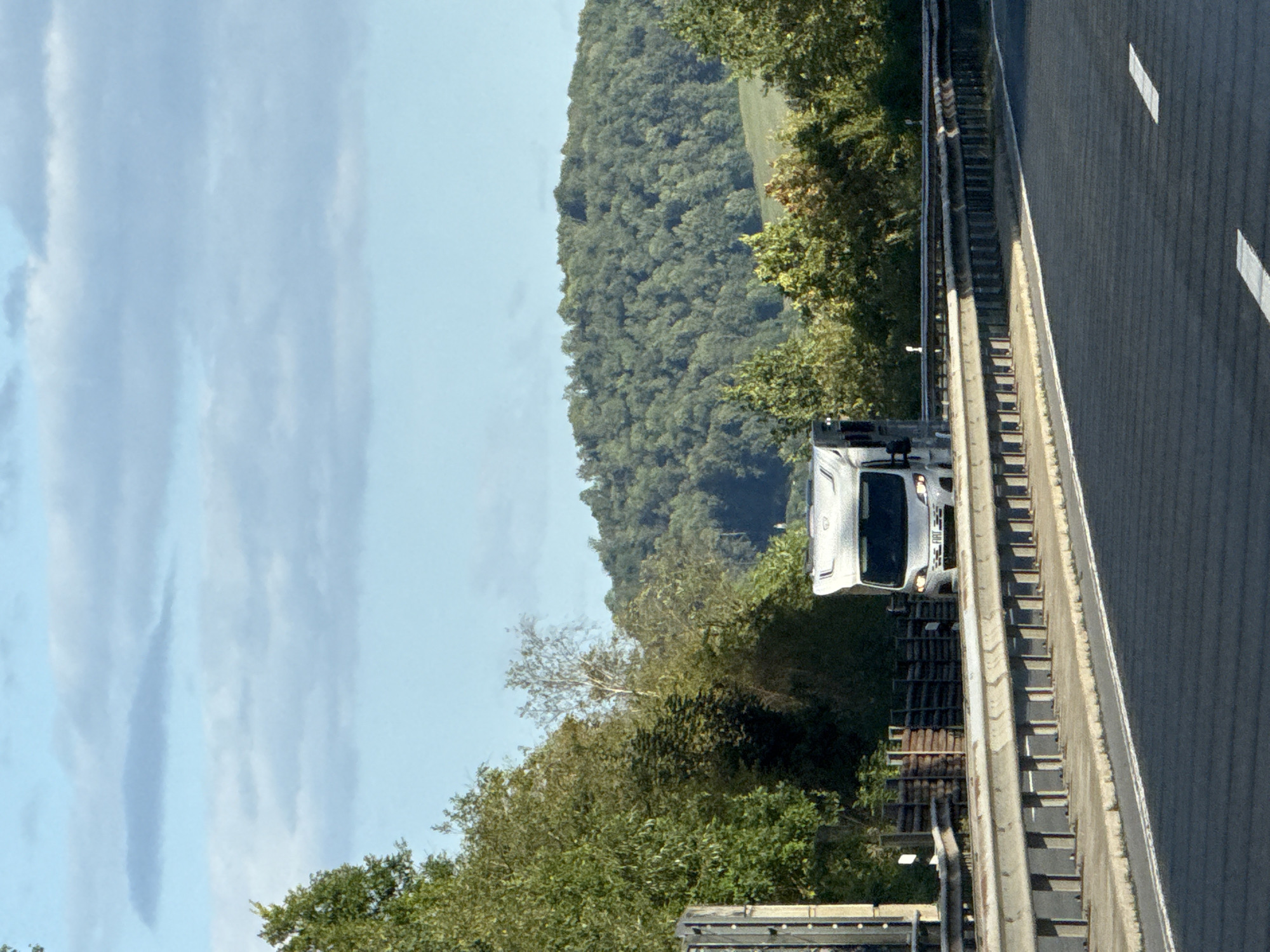 A white camper van is traveling on a multi-lane road in Herzberg, Germany. The road has asphalt paving and white lane markings. A metal guardrail runs along the right side of the road. On the left side of the road, there is a concrete barrier. Beyond the road and guardrail, there are trees and a gently rolling, forested hill. The sky is blue with scattered clouds. The camper van appears to be the only vehicle visible on the road. The image seems to have been taken from a moving vehicle.