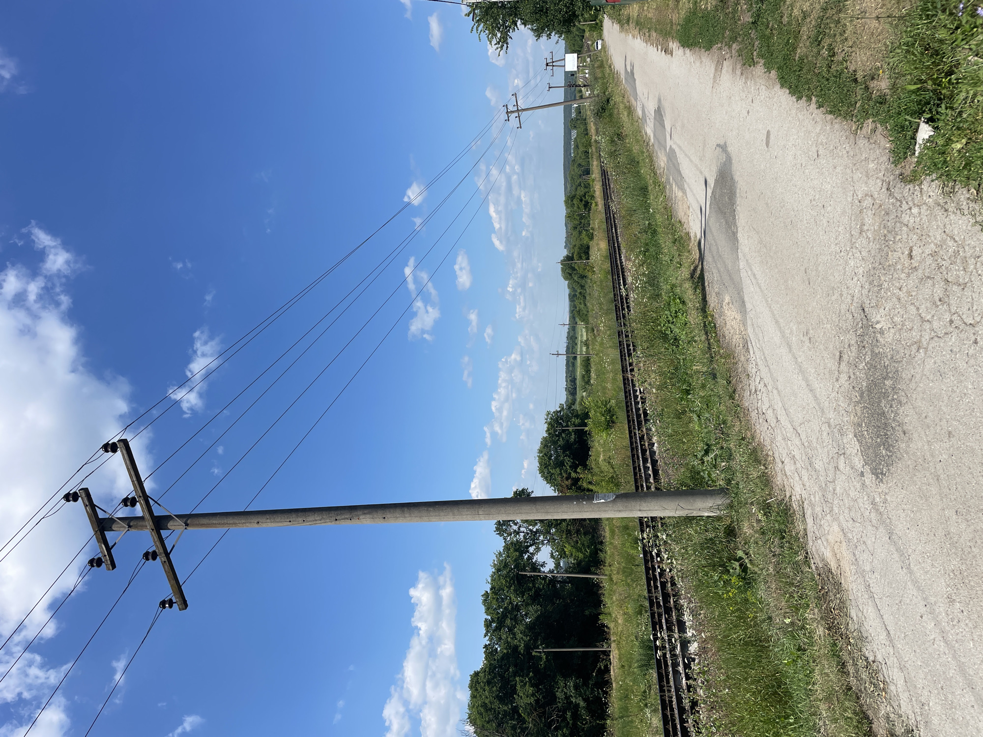 A dirt road runs alongside railroad tracks under a bright blue sky with scattered white clouds. A concrete utility pole with multiple wires stands on the left side of the road. The surrounding landscape is green and rural, with trees and fields visible in the distance. The scene appears to be in the village of Barsuki, Russia.