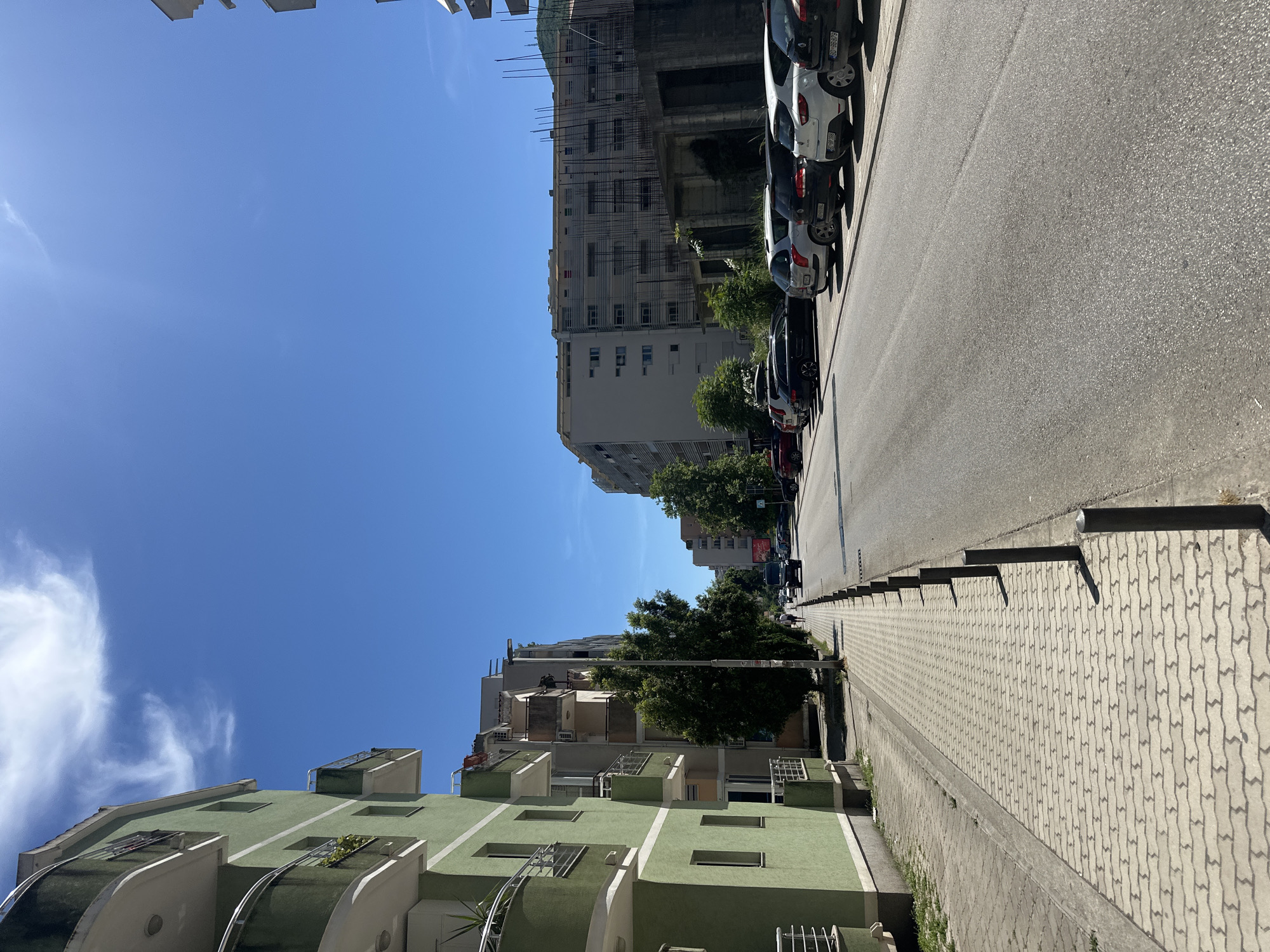 The image depicts a sunlit street in Budva, Montenegro. On the left, a light green apartment building with balconies lines the sidewalk, which is paved with interlocking concrete pavers and bordered by metal bollards. The street, asphalted and in good condition, runs towards the background, with several cars parked along the right side. Beyond the parked cars, a construction site is visible, featuring scaffolding and unfinished concrete structures adjacent to a multi-story building. Trees are sparsely planted along the sidewalk and in front of the buildings. The sky is a clear, bright blue with a few wispy white clouds. A black van is visible driving away from the camera on the street, and a person is walking on the sidewalk in the distance.