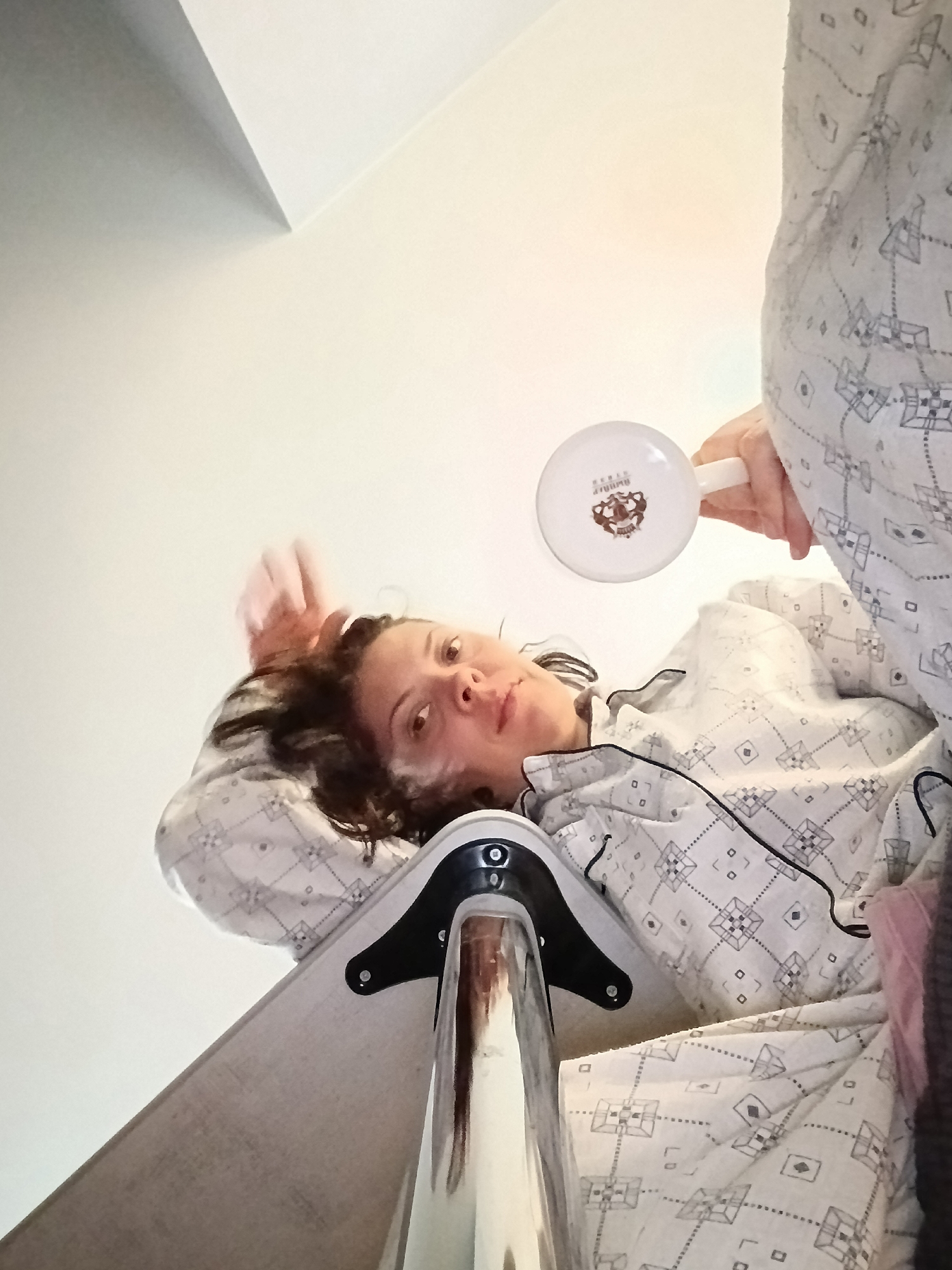 The image shows a low-angle, upward shot of a woman in pajamas holding a white mug with a logo. She is lying down, possibly in bed or on a sofa, with her head tilted back and one arm raised. The background is a plain wall. The woman appears to be in a domestic setting. There is no identifiable information in the image that would place the location in Saratov, Russia.