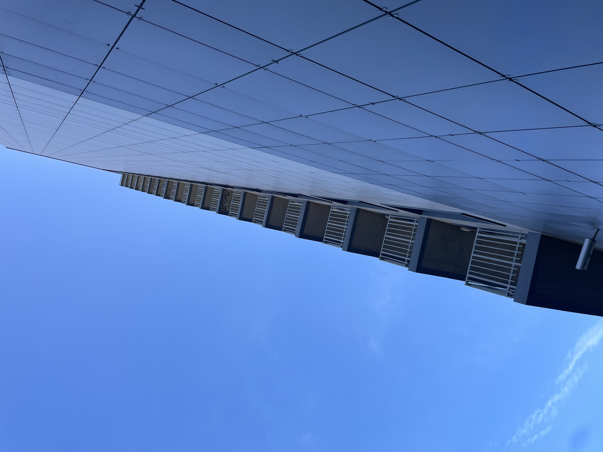 An exterior, low-angle, full shot captures a modern building with a blue glass facade and a concrete and white-railed balcony extending upwards along the left side. The sky is a clear, bright blue with a few wispy white clouds visible near the bottom left. The building's facade is composed of numerous rectangular panels, reflecting the sky and creating a sleek, contemporary look. The balconies are stacked vertically, each with a dark concrete base and white railings. The image appears to be taken in an urban environment, given the architecture of the building.