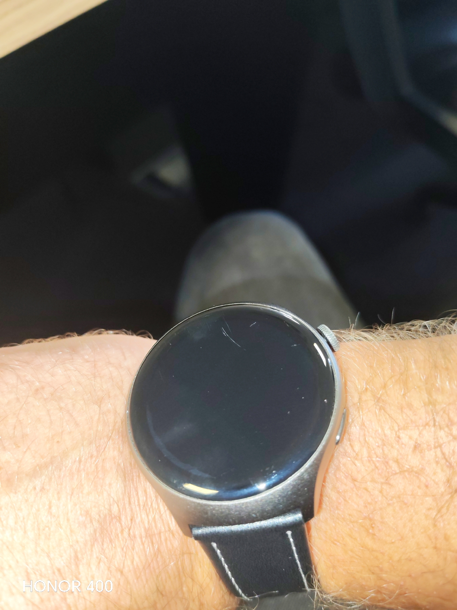 A close-up, waist-up shot shows a person wearing a silver smartwatch with a black band on their left wrist. The watch face is dark and reflective, showing some light scratches. The person's arm is covered in light hair. In the background, out of focus, is a dark environment that appears to be the interior of a vehicle, possibly a car, with a hint of a person's leg wearing dark pants. The lighting suggests an indoor setting or a dimly lit area. There are no identifiable landmarks or context to suggest a specific location in Baku, Azerbaijan.