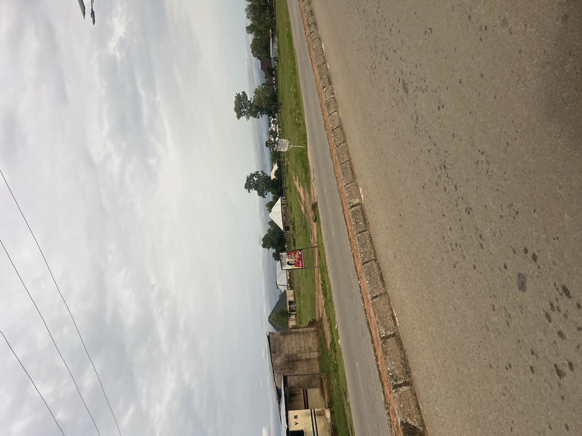 The image depicts a roadside scene in Jalingo, Nigeria. The perspective is from street level, looking upwards and sideways due to the orientation of the camera. On the right side of the image is a paved road with visible stains and wear. The road is bordered by a concrete curb and a strip of dirt and grass.
To the left of the road, there is a grassy area with several structures. One prominent feature is a large billboard with a portrait of a man and some text, although the text is largely unreadable. Behind the billboard, there are some trees, and further back, more buildings and vegetation can be seen. There's a white tent-like structure visible in the middle ground. In the foreground on the left, power lines stretch across the sky, indicating the presence of electricity infrastructure. The sky is overcast with clouds, suggesting it is daytime, though the light is diffused. The overall scene appears to be a somewhat rural or semi-urban area. No specific activity or event is evident, and no clear emotions are conveyed.
