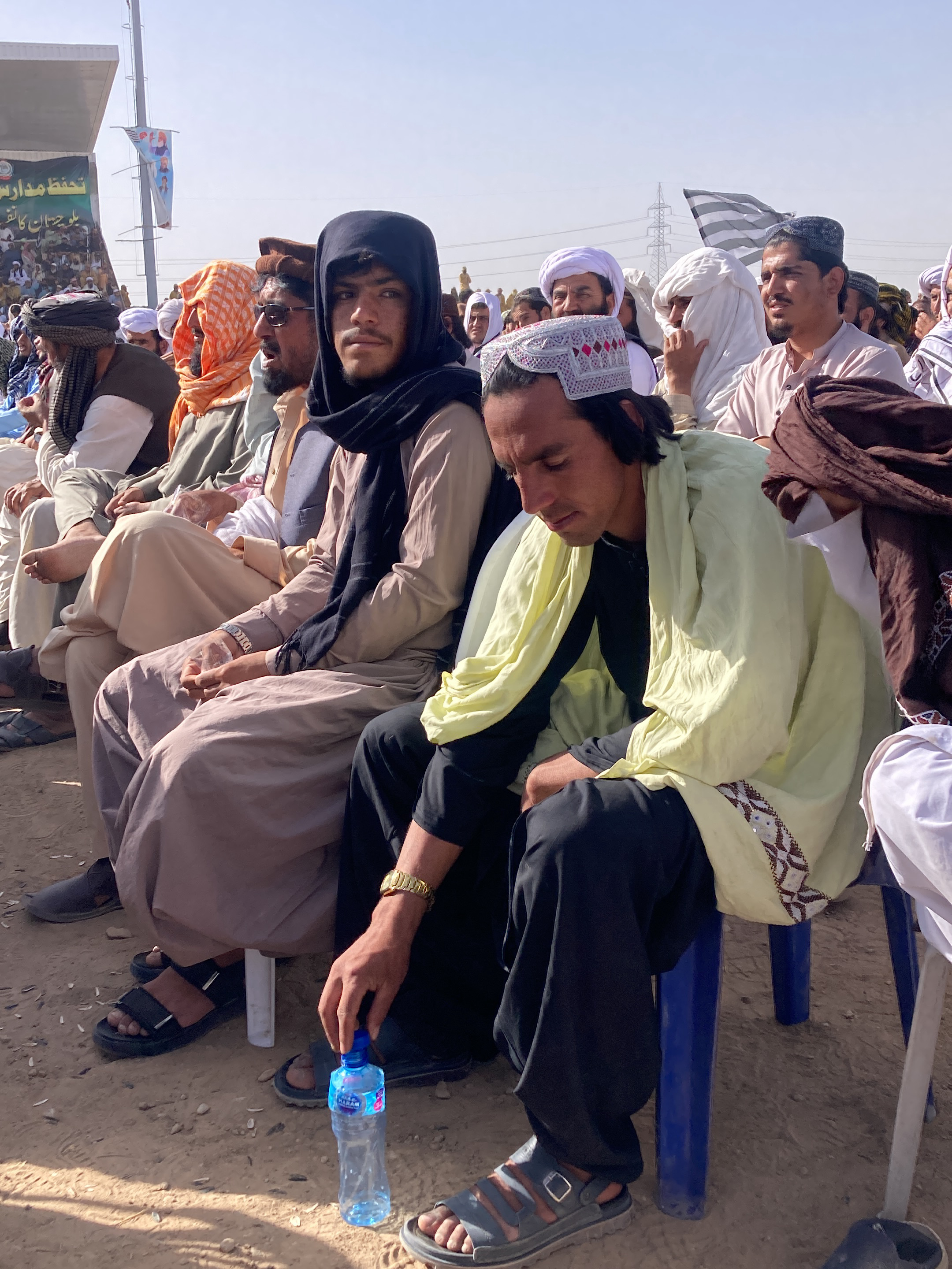 This image captures an outdoor gathering of men in Pishin, Pakistan, under a clear, sunny sky. A group of men, dressed in traditional South Asian attire including shalwar kameez, turbans, and various head coverings, are seated on blue plastic chairs on a dusty, bare ground surface.
In the foreground, a man with long dark hair wears a white embroidered cap, a light yellow shawl draped over a dark top, and dark shalwar. He has a gold-colored watch on his left wrist and is reaching down for a clear plastic water bottle placed on the ground between his feet. To his left, another man, central to the frame, wears a black head covering or shawl over light-colored traditional clothing, sporting a beard and mustache, and looking straight ahead with a calm expression. Further left, another individual wears an orange head covering and glasses. Most of the men are wearing sandals.
In the background, on the left, a large banner is partially visible. It features text in green and red script, likely Urdu or Pashto. Discernible phrases include "حفظ مدارس" (protection/preservation of religious schools/seminaries), and the word "طالبان" (Taliban). Other visible text fragments appear to relate to "امن" (peace) and "استحکام" (stability). On the far right in the background, a black and white striped flag is also visible.
The scene suggests a public event, possibly a rally or community meeting, taking place during the daytime. The men generally appear calm and observant.