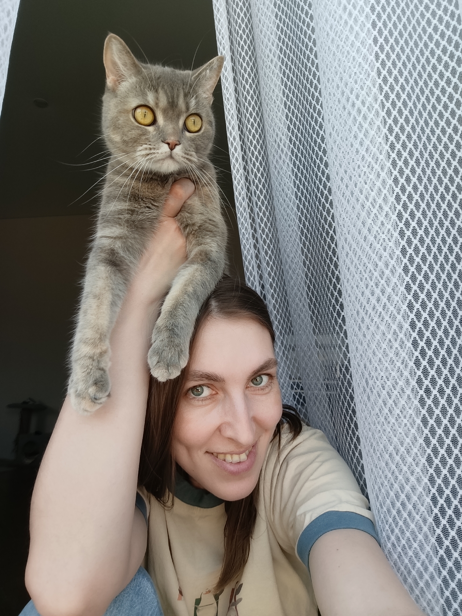 A woman is holding a gray tabby cat in front of a window covered with white mesh curtains. The woman is smiling and looking at the camera. The cat is also looking at the camera with wide yellow eyes. The woman is wearing a beige t-shirt with blue trim. The scene is indoors. There is no indication of being located in Chaikovsky, Russia.