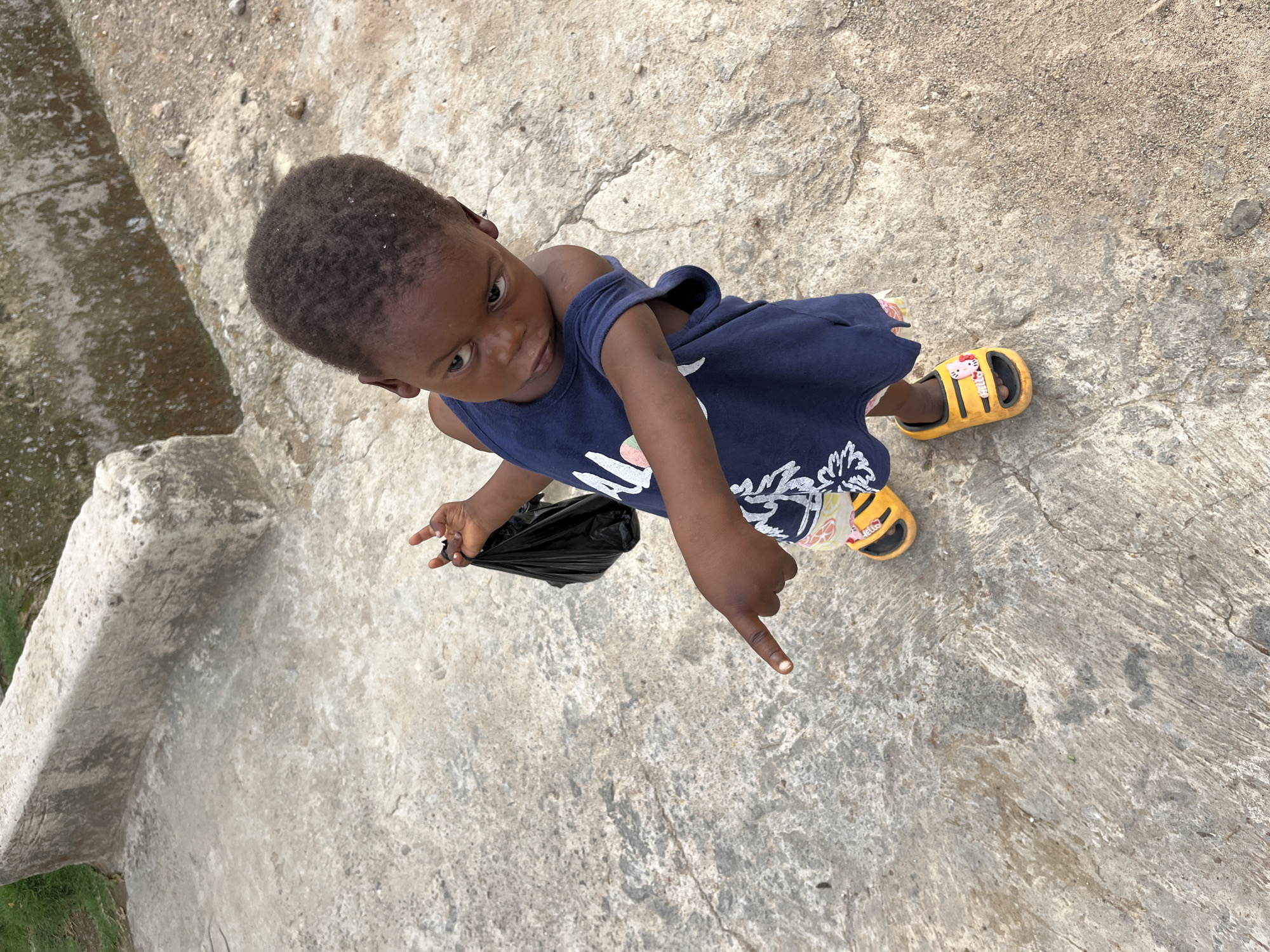 A young child stands on a concrete surface. The child is Black, has short, dark hair, and wears a navy-blue dress with light-colored floral patterns and yellow sandals. The child is holding a black object, likely a bag, in one hand, and pointing with the index finger of the other hand toward the ground. The background and surrounding area appear to be concrete, suggesting an urban environment in Abuja, Nigeria.