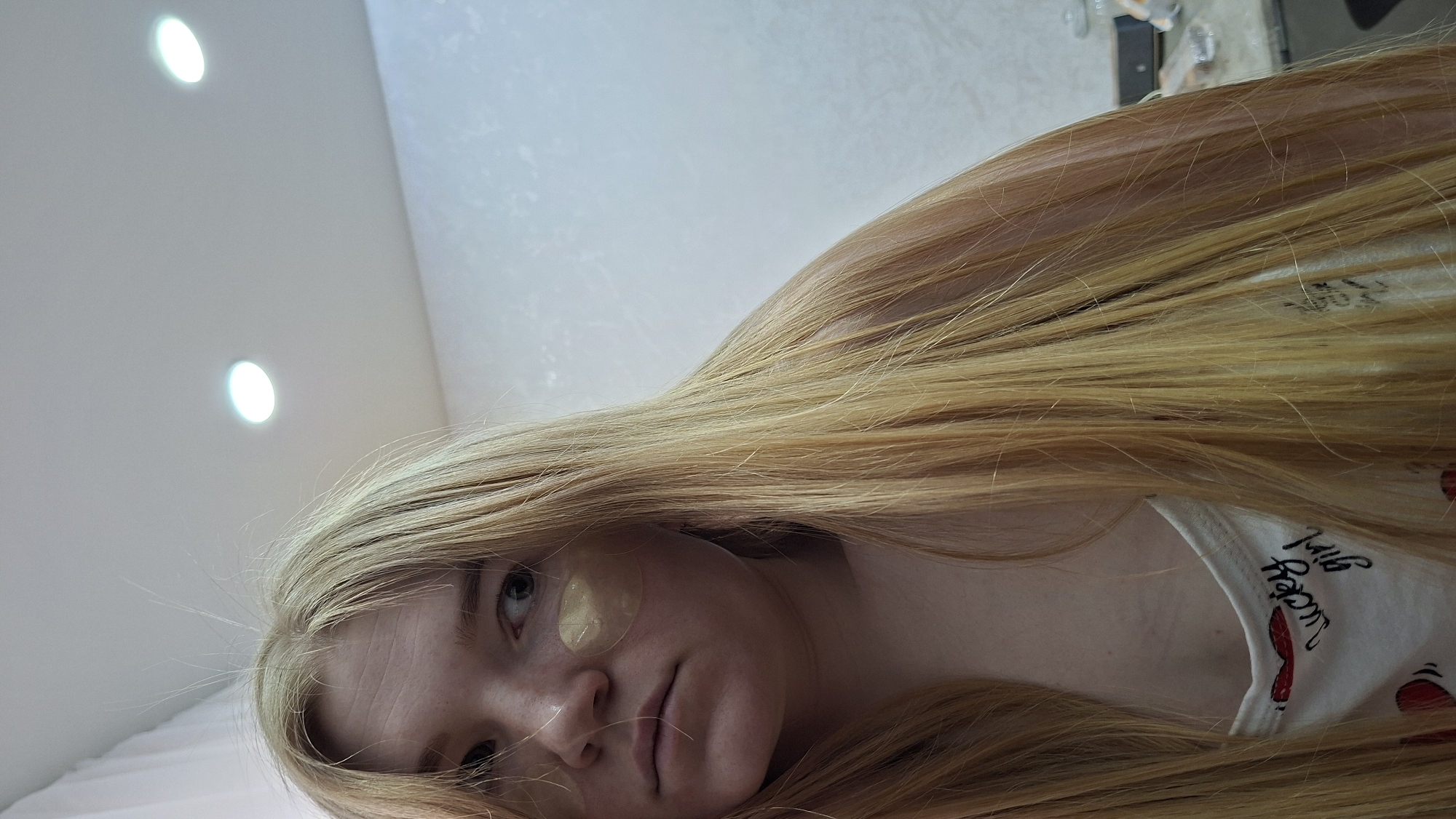 A young person with long, straight blonde hair lies or reclines in an indoor setting. The image is taken from a low angle, looking upwards towards the subject and a white ceiling with two bright recessed lights. The person's face is partially visible, oriented sideways in the frame, with their expression appearing neutral or slightly pensive. Gold-colored hydrogel eye patches are visible beneath both eyes. The subject is wearing a white garment, possibly a t-shirt or tank top, which has text and a graphic. The text "Lucky girl" is clearly visible in a cursive-style font, alongside what appears to be part of a red lips or heart graphic. The long blonde hair is spread out, covering parts of the garment and extending across the frame. The overall scene suggests a moment of relaxation or self-care. The lighting is artificial, indicative of an indoor environment.