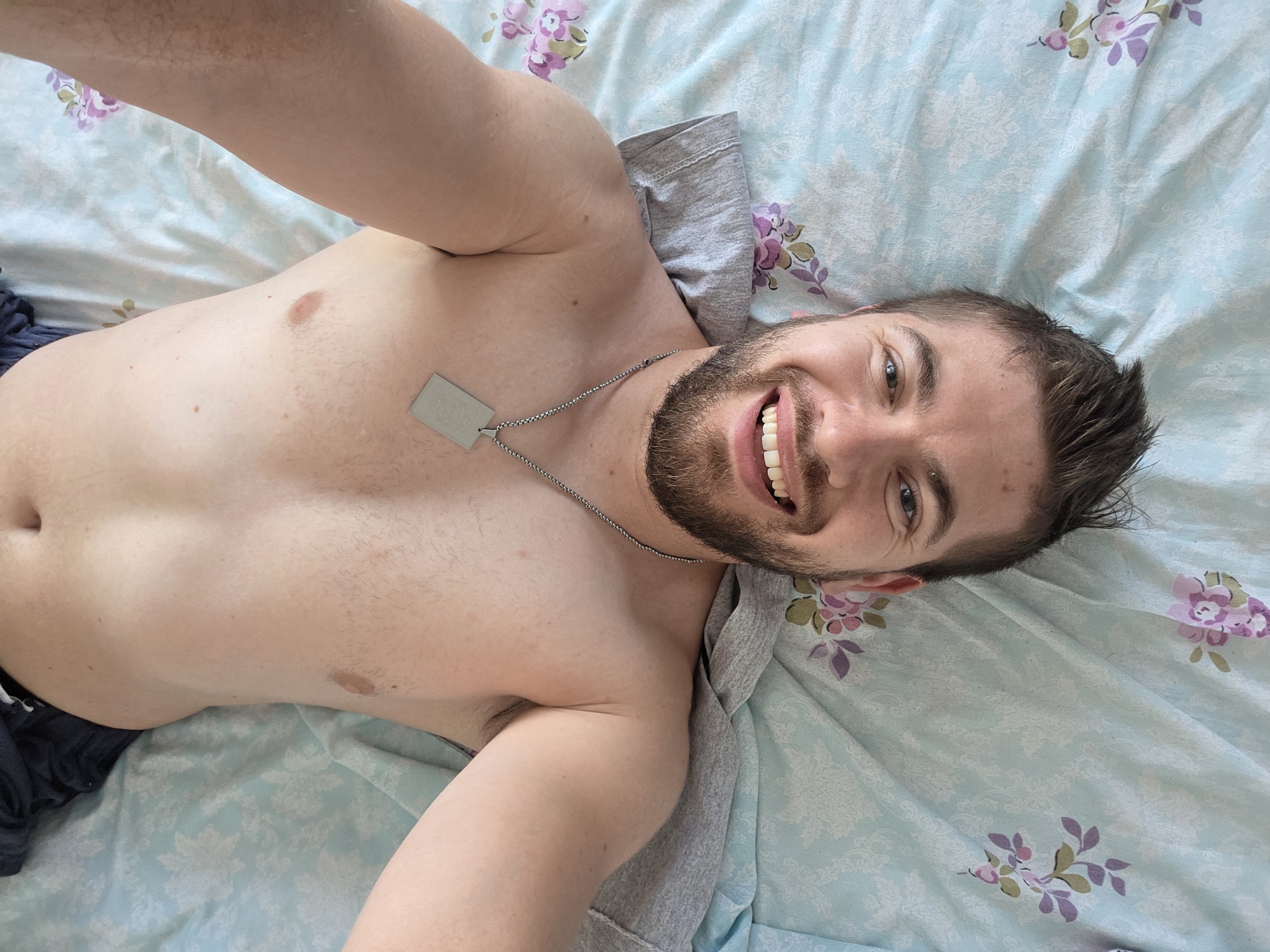 A man with a beard and styled hair is lying on a bed with a blue floral patterned sheet. He is wearing a silver necklace with a rectangular pendant. The man is shirtless and appears to be taking a selfie. The scene is an indoor environment, likely a bedroom. The location of Yagodovo, Bulgaria is not depicted or suggested by the image.