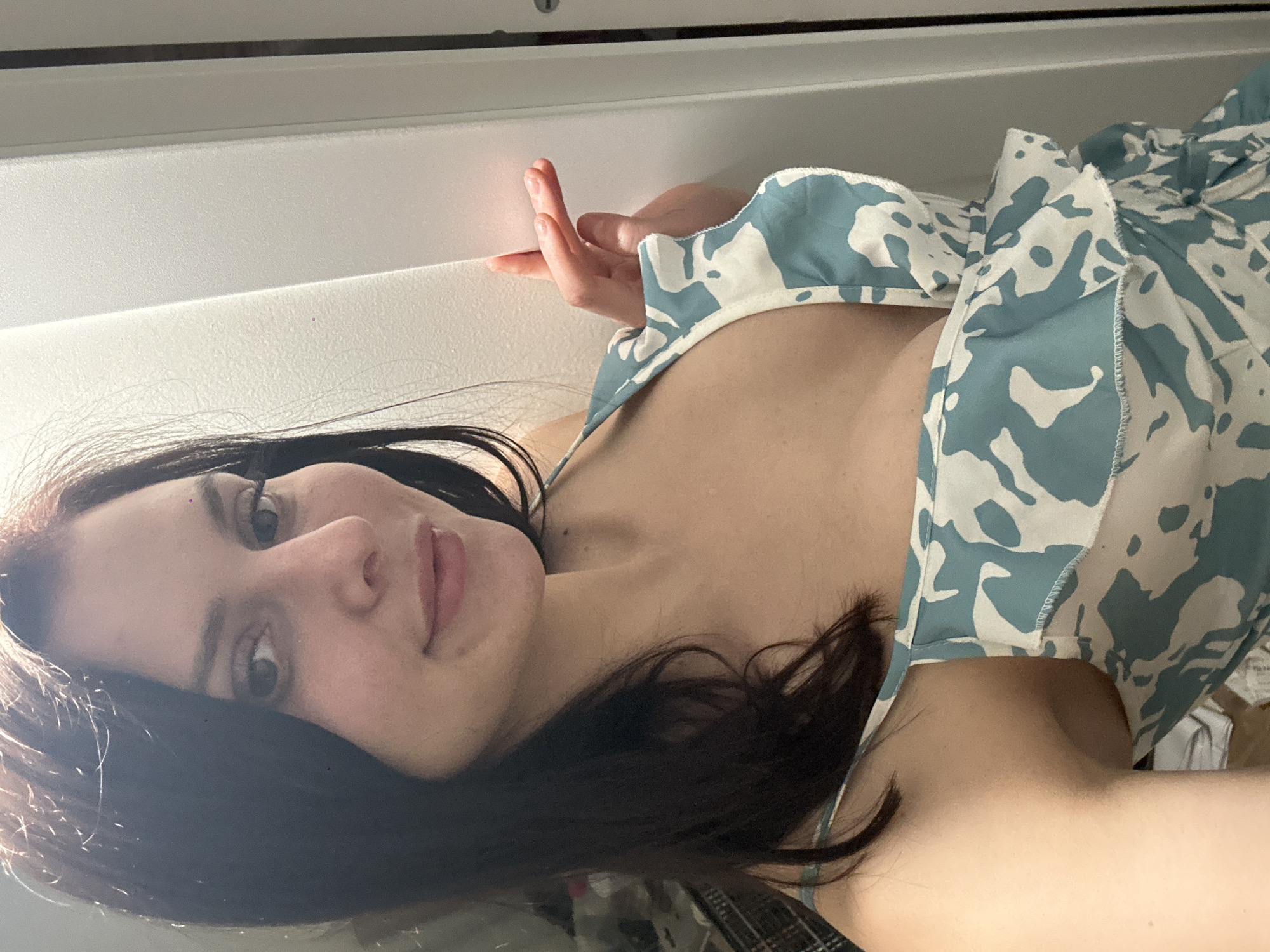 The image shows a close-up, overhead shot of a woman's face and upper body, oriented sideways. She has dark hair that falls around her face and shoulders. Her eyes are open and she is looking directly at the camera with a neutral expression. Her lips are slightly parted. She is wearing a light-colored garment with a blue and white abstract pattern. Her right hand is visible, resting near her face, with her fingers slightly curled. The background appears to be a neutral, textured surface, possibly a wall or ceiling, with a horizontal line across the top of the image. The lighting suggests it could be daytime, with light coming from the left, creating soft shadows. There is no visible text in the image. The overall impression is that of a casual, personal photograph.