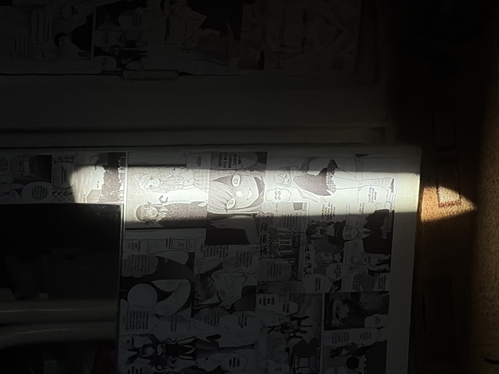 The image shows a wall covered with manga panels. A shaft of light illuminates the lower portion of the wall and a small section of the floor, casting a geometric shadow. The manga panels depict various characters and scenes, including anime-style figures with large eyes and speech bubbles, suggesting a narrative. The overall lighting suggests it is daytime and the scene is indoors, with natural light entering the space. The environment appears to be a domestic or personal space given the decoration.