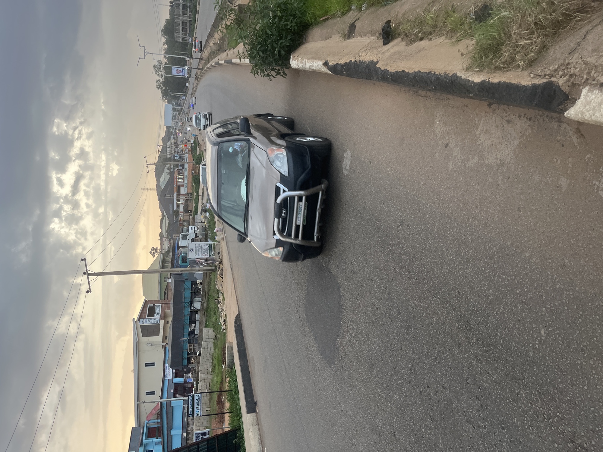 The image shows a grey SUV driving on a paved road in Gura Top, Nigeria. The SUV is in the foreground, with its front end facing left and slightly towards the camera. It has a silver bull bar attached to the front bumper. The road is asphalt and appears to be a multi-lane highway. On the right side of the road, there is a concrete barrier with some overgrown grass and dirt. In the background, there are several buildings, some of which appear to be commercial establishments with signage. Power lines and streetlights are visible overhead. The sky is overcast with a mix of grey and white clouds, suggesting it might be evening or an overcast day. Other vehicles are visible further down the road, indicating ongoing traffic.