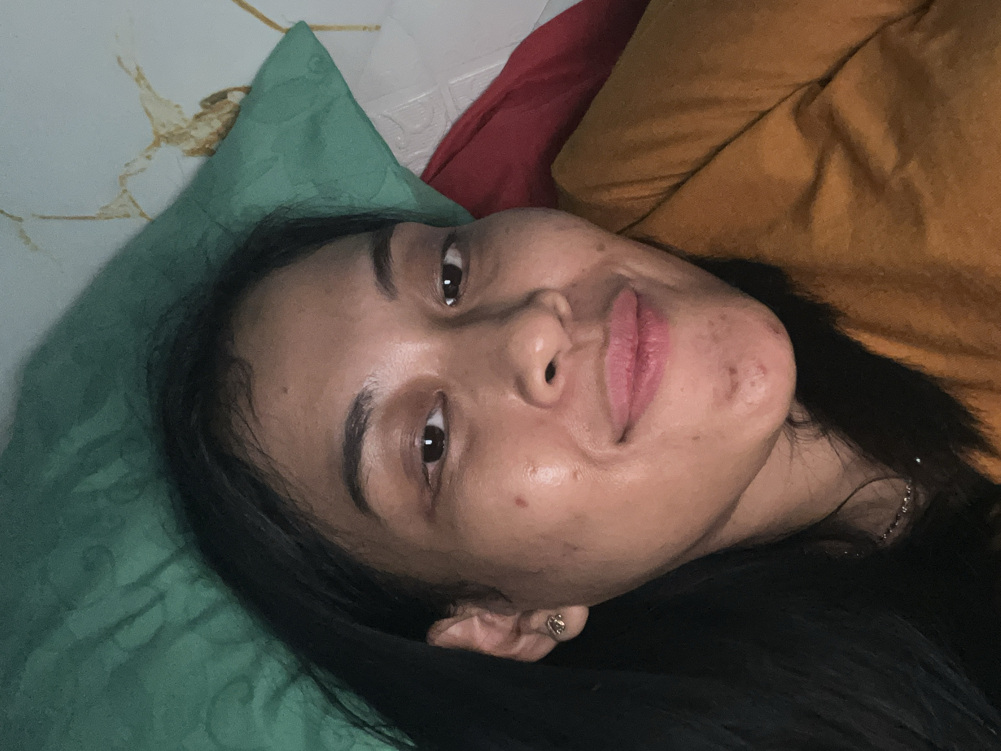 The image is a close-up selfie of a young woman lying down, possibly in bed. She has dark hair and is wearing an orange garment. Her face is turned towards the camera, and she has a slight smile. The background consists of pillows and bedding, with one pillow appearing green and another red. The lighting is dim. The image does not contain any information or visual cues that would indicate its location in Kecamatan Pati, Indonesia.