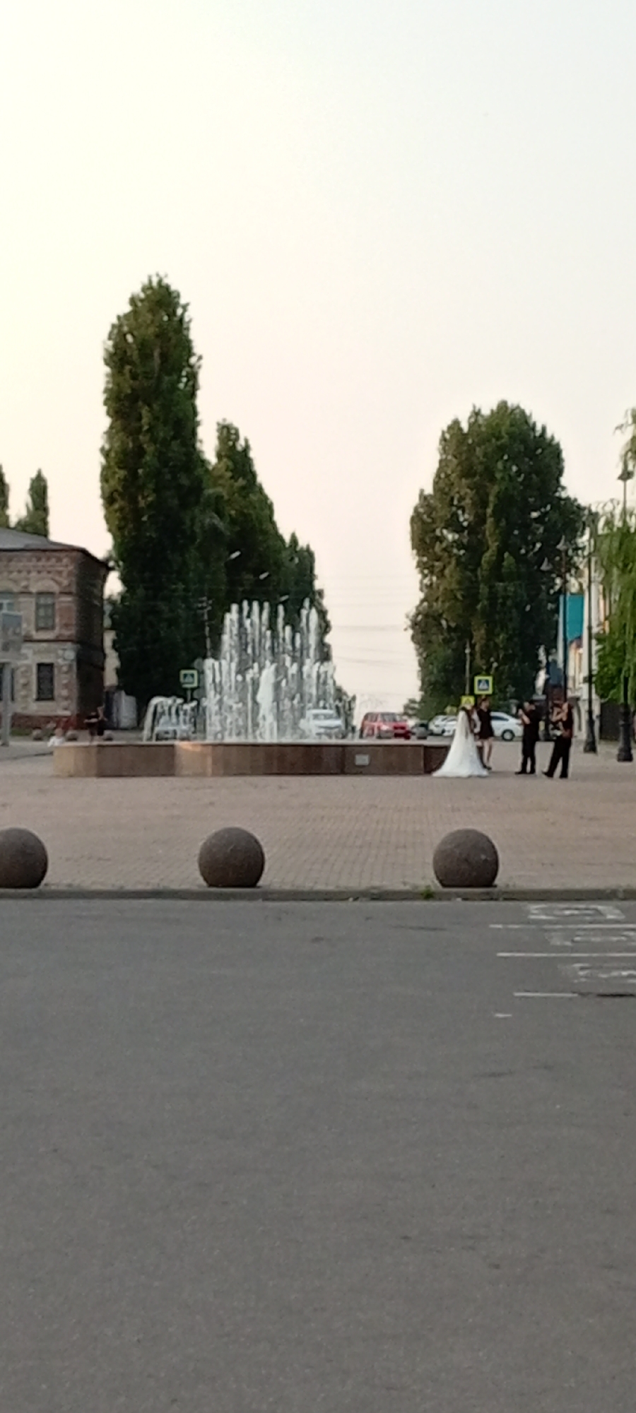 In Kirsanov, Russia, a street scene unfolds with a large public fountain as the central feature. The fountain, with multiple jets of water, is situated in a paved plaza. Surrounding the plaza are trees, a brick building on the left, and various vehicles, including cars, in the background. In the foreground, a street with a dashed white line and what appear to be bollards are visible. A group of people are gathered near the fountain, including what appears to be a bride in a white dress and other individuals dressed in dark clothing, possibly a wedding party. One person is holding a phone, seemingly taking a photo or video. The sky is a pale, overcast color, suggesting either early morning or late afternoon.