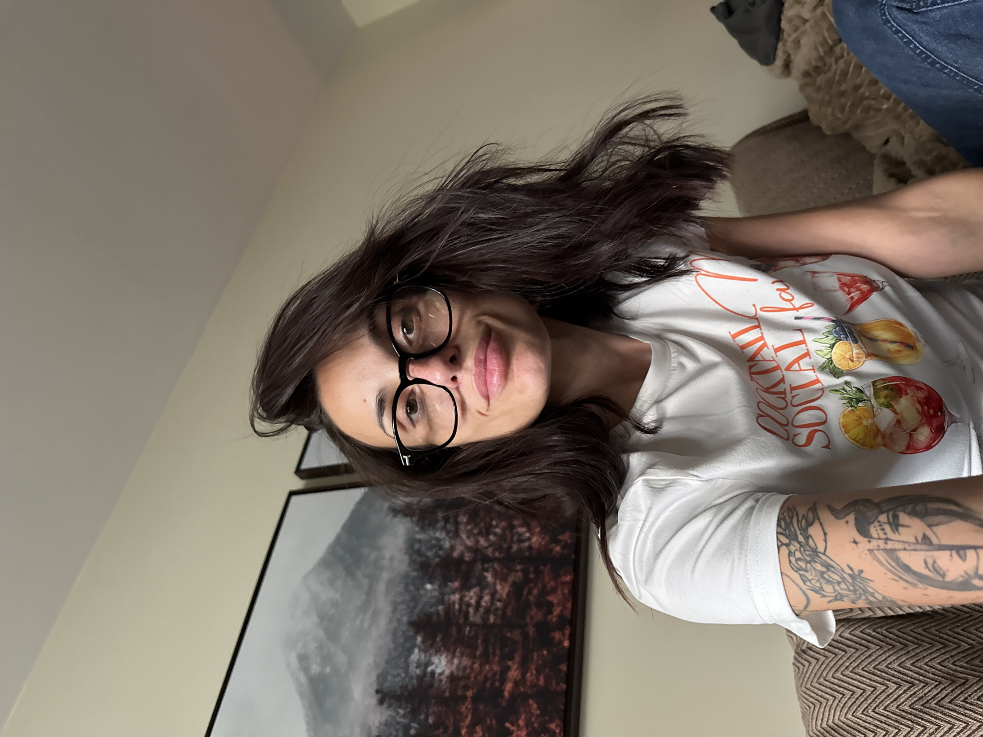 A woman with dark hair and glasses is looking at the camera. She is wearing a white t-shirt with a graphic print. Her left arm has a tattoo. There is a framed picture behind her and a couch or seat to her right. The location is unlikely to be Pleven, Bulgaria.