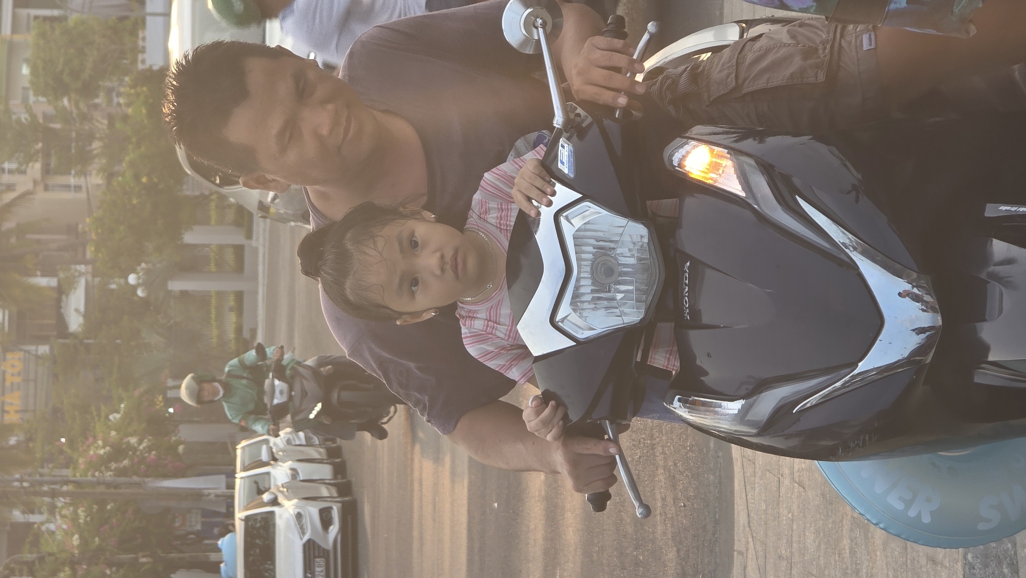 An adult male and a young child are seated on a black Honda scooter, captured in a vertical orientation. The adult, with short dark hair and wearing a dark t-shirt, is positioned behind the child, holding the scooter's handlebars. The child, with dark hair pulled back and wearing a pink and white striped t-shirt, sits in front of the adult, also gripping the handlebars. She looks directly at the camera with a neutral expression, wearing small earrings and a necklace.
The scooter is black with silver and white detailing on the front. The "HONDA" logo is visible on its front panel, and one of its indicator lights is illuminated. The scene appears to be an outdoor setting, likely a street or road in Nha Trang, Vietnam, bathed in the warm, soft light of the late afternoon or golden hour.
In the background, a paved road stretches out, lined with trees and buildings. Another person on a scooter, wearing a green jacket and a helmet, is visible further down the road, and several white cars are also present. A blue and white inflatable inner tube, partially displaying the text "NER SWI" (likely part of "POWER SWIM" or similar), is visible in the bottom right corner of the image. On a building in the distant background, the text "HATÔI" can be discerned. The overall impression is a casual, everyday scene.