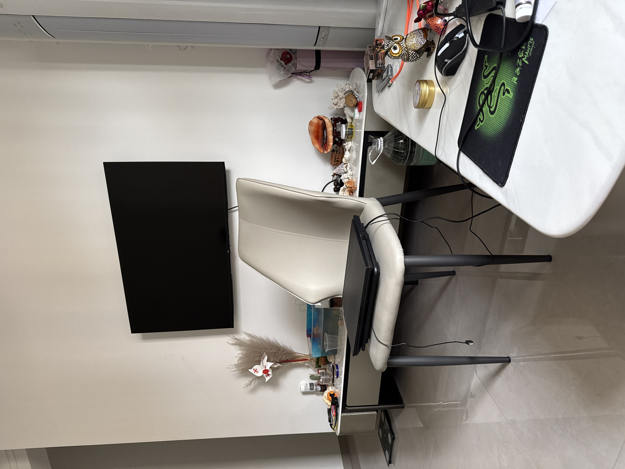 The image depicts an indoor living space. A television is mounted on a white wall. In front of the television, a light-colored chair is positioned, with a closed black laptop resting on its seat. A low, white entertainment unit is visible beneath the television, displaying various items including a decorative dried floral arrangement, a glass of water, and what appears to be a small aquarium. To the right of the chair, a white side table holds several decorative seashells, a large conch shell, and a small jar. Further to the right, a tall, white vertical appliance, possibly an air conditioner, stands against the wall. In the foreground, a portion of a white marble-topped table is visible, featuring a computer mouse, a mousepad with a green logo, and a small golden tin. The floor is tiled with a light-colored, reflective material. The scene is lit by artificial indoor lighting. The presence of the entertainment unit, television, and seating suggests a living room or lounge area. The overall impression is one of a tidy, domestic environment. The image does not provide any explicit visual cues that would allow for its specific location within the city of Huizhou, China.