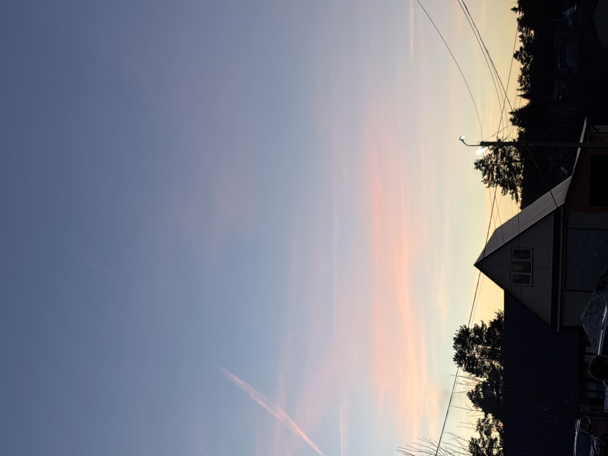 The image shows a twilight or dawn sky with pastel colors of blue, pink, and yellow. A contrail is visible in the lower-left portion of the sky. In the lower right, there is a house with an A-frame roof and visible windows. Silhouetted trees are next to the house. Power lines extend across the sky from the upper right. A street light is next to the house. The location is Chaikovsky, Russia.