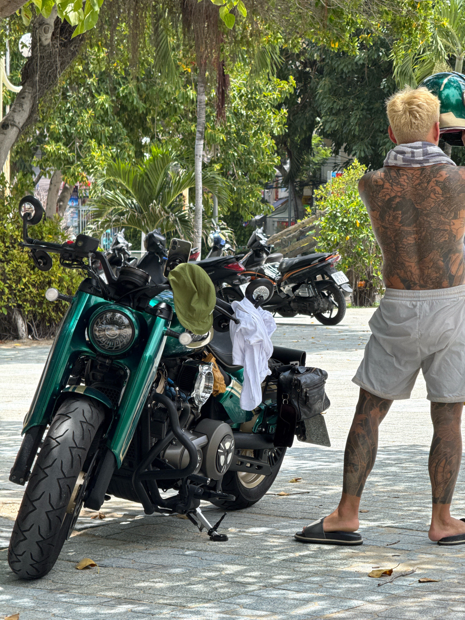A man with extensive black tattoos covering his back and legs stands next to a green motorcycle in Nha Trang, Vietnam. He has bleached blonde hair and is wearing a grey and white checkered towel around his neck, light grey shorts, and black slide sandals. The motorcycle is parked on a paved walkway under trees with dappled sunlight. A green cap is hanging from the handlebars, and a white shirt is draped over the seat. In the background, other motorcycles are visible, and beyond that, trees and a building with a fence. The scene appears to be outdoors, possibly in a public area or parking lot.