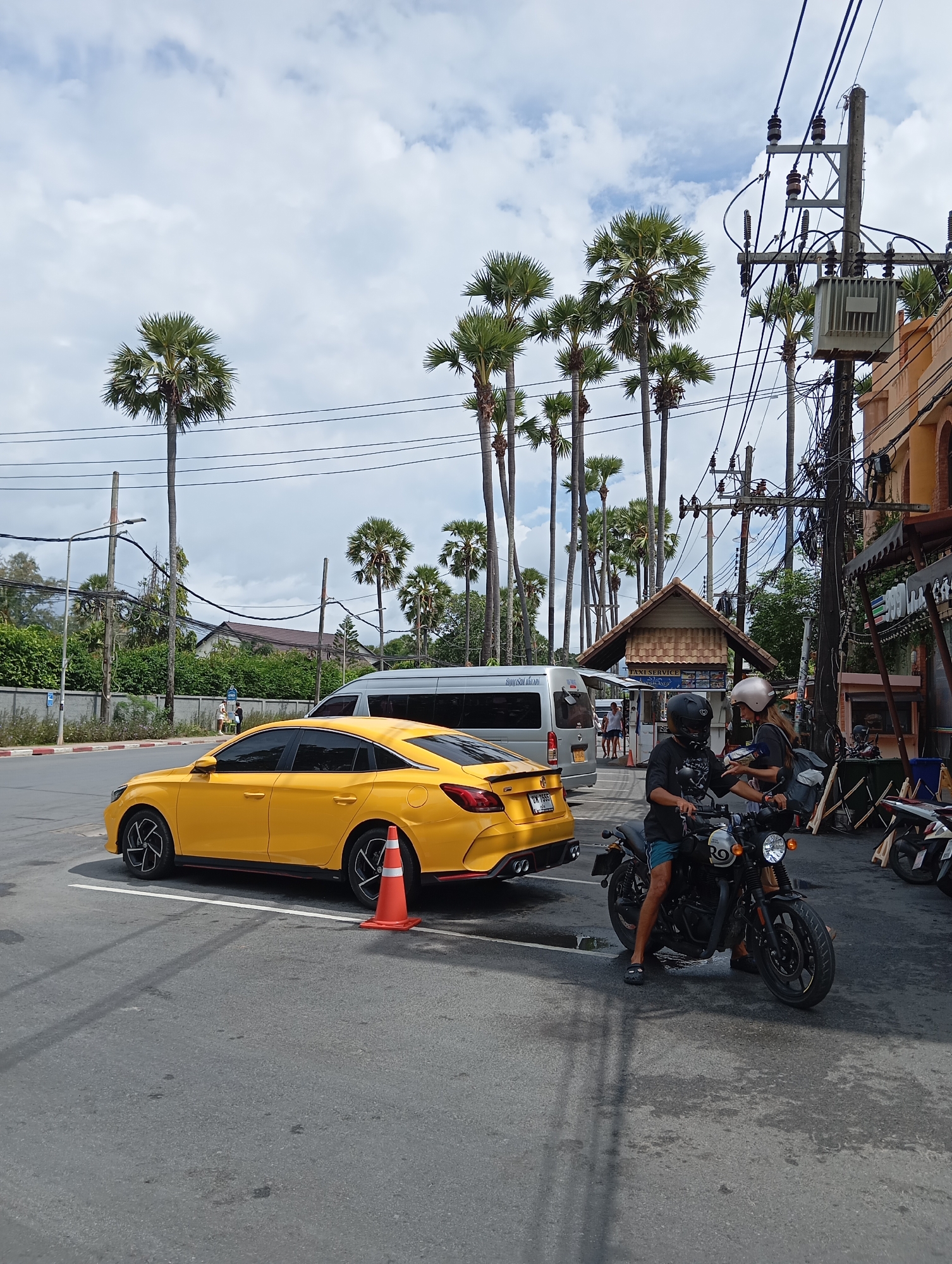 The image depicts a street scene in an outdoor urban environment. In the foreground, a yellow sedan is parked to the left, facing right, with an orange traffic cone near its rear. To the right of the car, a person is sitting on a black motorcycle. The person is wearing a black t-shirt, blue shorts, sandals, and a black helmet. They are interacting with another person, who is also on the motorcycle and wearing a light-colored helmet and a dark top.
In the background, a silver van is parked. Several tall palm trees are visible throughout the scene, suggesting a tropical or subtropical location. A building with a tiled roof, possibly a taxi service, is situated behind the van. Overhead, a complex network of electrical wires and utility poles is prominent. The sky is partially cloudy. The overall scene is a snapshot of a typical day on a street, with vehicles, people, and infrastructure present.
