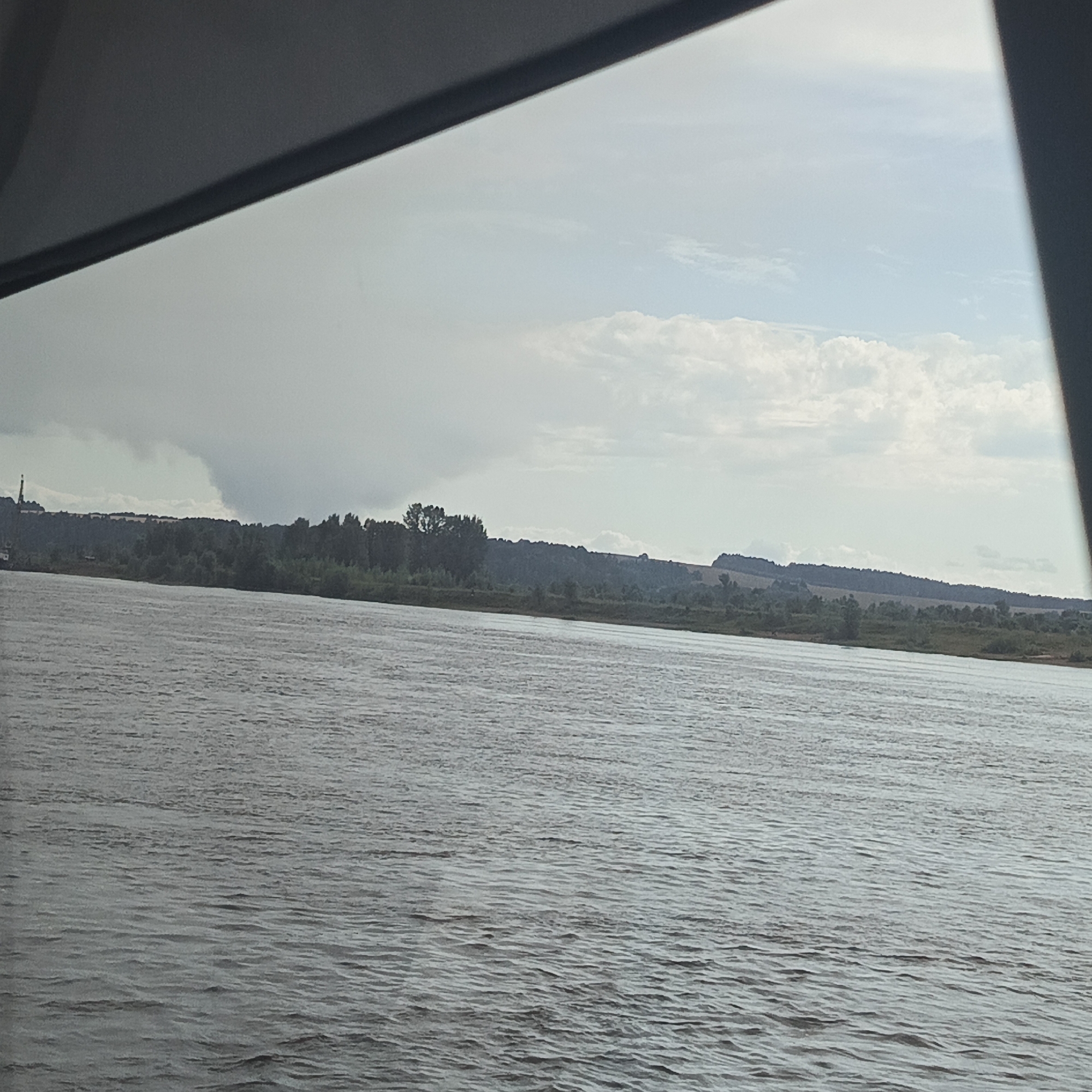 The image shows a wide, murky river flowing through a natural landscape under a cloudy sky. The river occupies the foreground, its surface rippled by a gentle breeze. In the background, a densely forested embankment stretches across the horizon, interrupted by patches of lighter vegetation and the rolling contours of hills. On the far left of the embankment, a tall industrial structure, possibly a smokestack or tower, is visible. The sky is overcast with a mix of grey clouds and brighter, sunlit areas, suggesting a dynamic weather system. The scene appears to be located near Nechkino, Russia, a town situated along the Votka River.