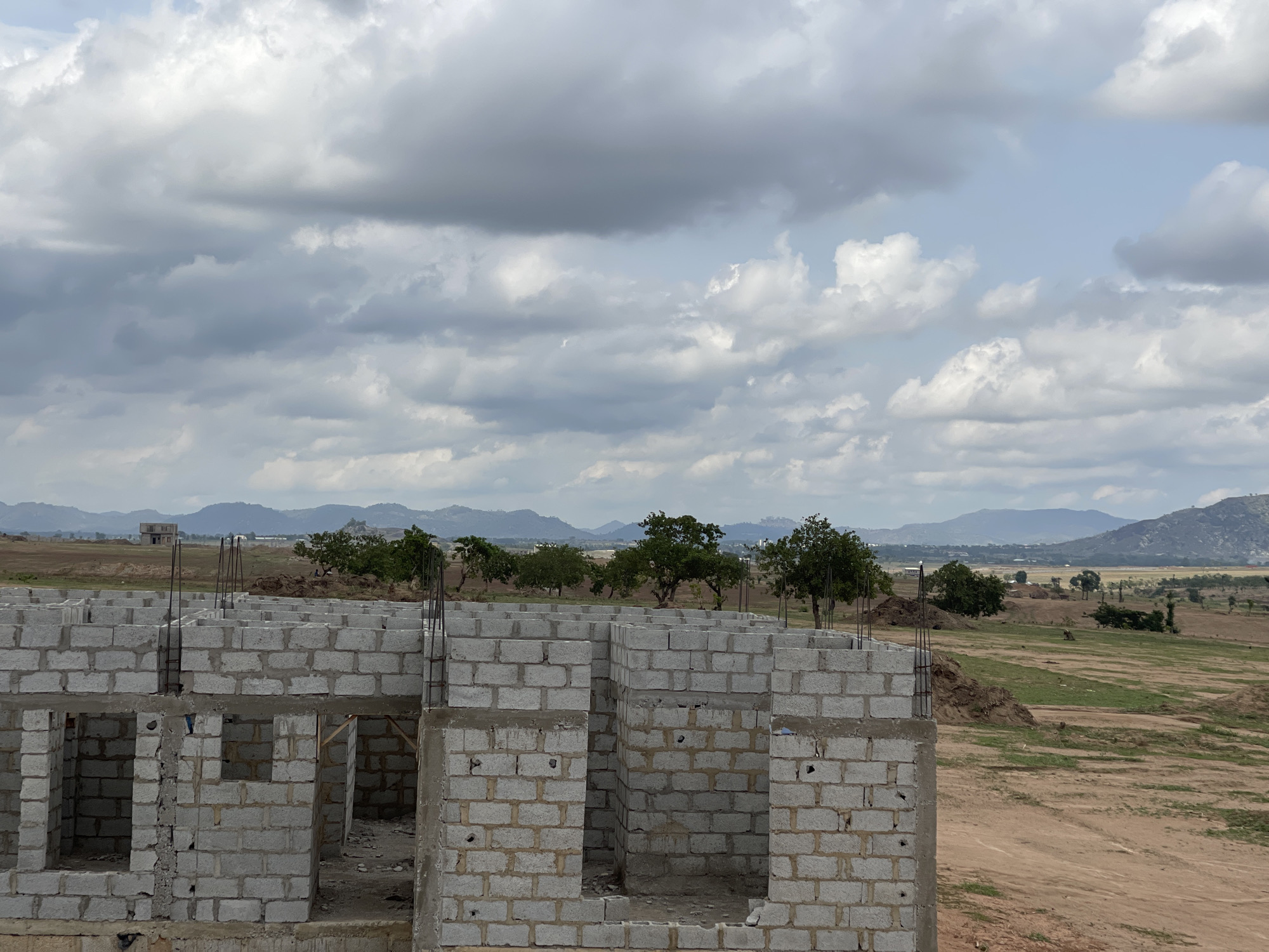 The image shows a partially constructed building in Angwan Sayawa, Nigeria. The building is made of grey concrete blocks and features steel reinforcement rods. The surrounding environment includes a grassy field, scattered trees, and hills in the distance. The sky is overcast with grey clouds.