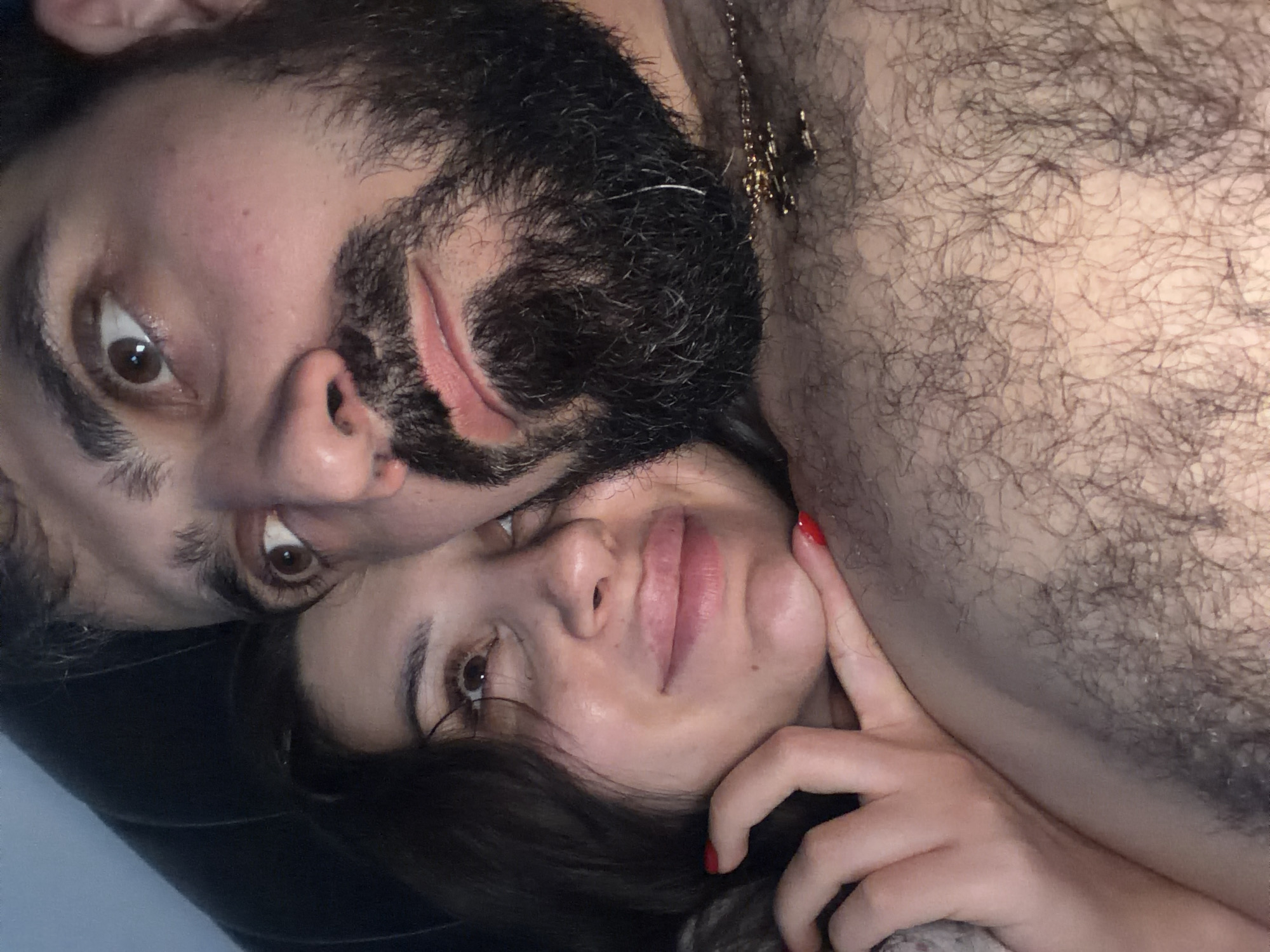 This close-up image captures a man and a woman lying down side-by-side, looking towards the camera. The setting appears to be indoors, likely a bed or couch, given the intimate and relaxed posture of the subjects and the soft lighting.
The man, positioned on the left and slightly above the woman, has dark hair and a full beard. His brown eyes are wide open, directed at the viewer. A significant portion of his chest and shoulder, covered in dark body hair, is visible on the right side of the frame. A gold-colored chain with a small pendant is visible around his neck, resting on his chest.
The woman is positioned below and to the right of the man. She has dark hair and brown eyes, also looking towards the camera with a calm expression. Her lips are slightly parted. Her right hand, adorned with bright red nail polish, gently touches her cheek, adding a touch of color to the scene.
The activity depicted is a shared, intimate moment between the two individuals. Their direct gazes suggest a connection with the camera or the person taking the photo. The emotions conveyed are generally relaxed and affectionate. There are no specific cues for the time of day or weather within the image. While the image itself does not show specific location details, the provided context indicates the location is Budva, Montenegro. There is no visible text in the media file.