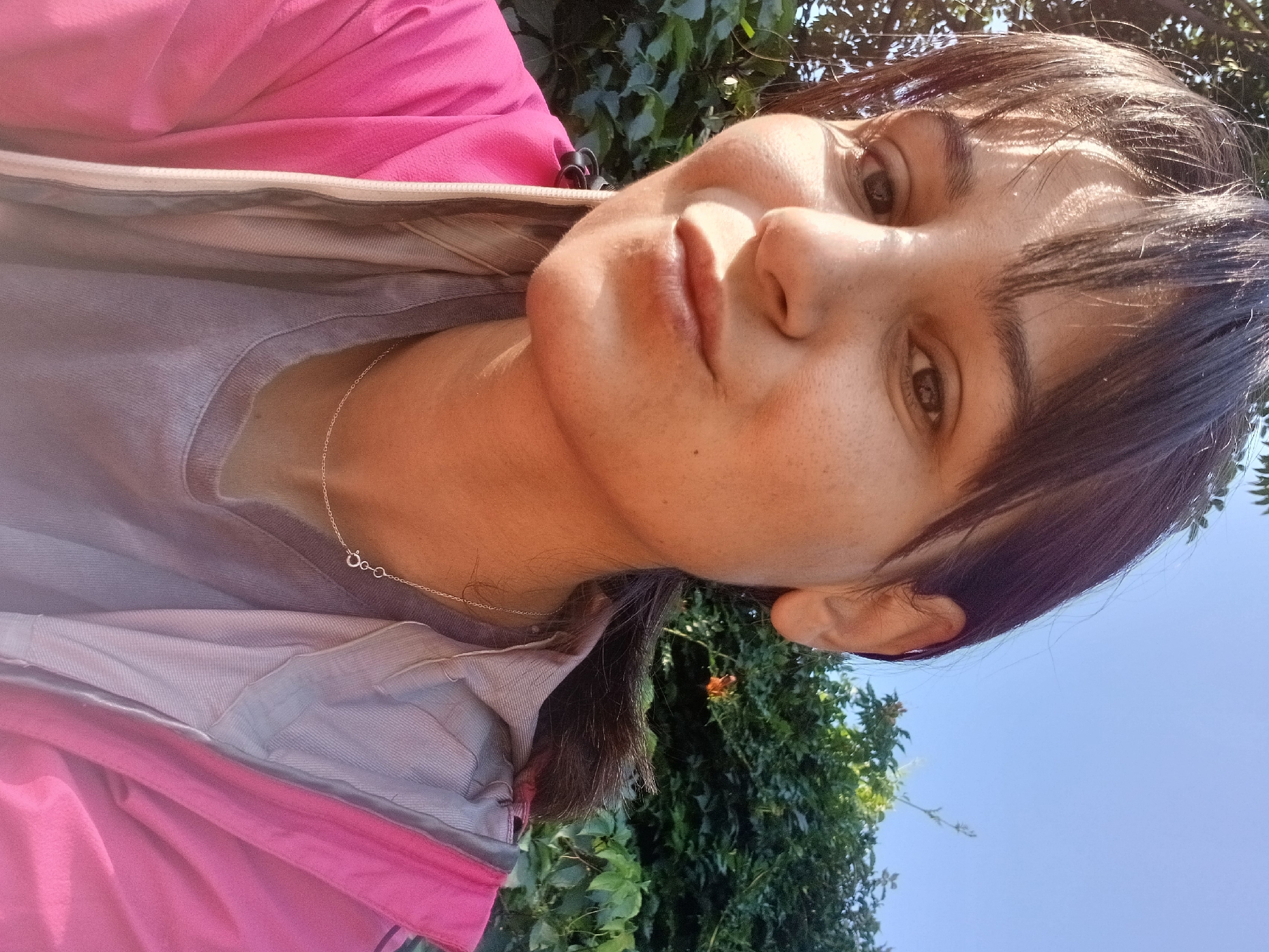 A young woman with dark hair and bangs smiles for a selfie outdoors, with lush green foliage and a bright blue sky behind her. She is wearing a pink jacket over a gray t-shirt and a delicate silver necklace. The sun casts a warm glow on her face, highlighting her features. The background suggests a natural, perhaps park-like, setting within a city.