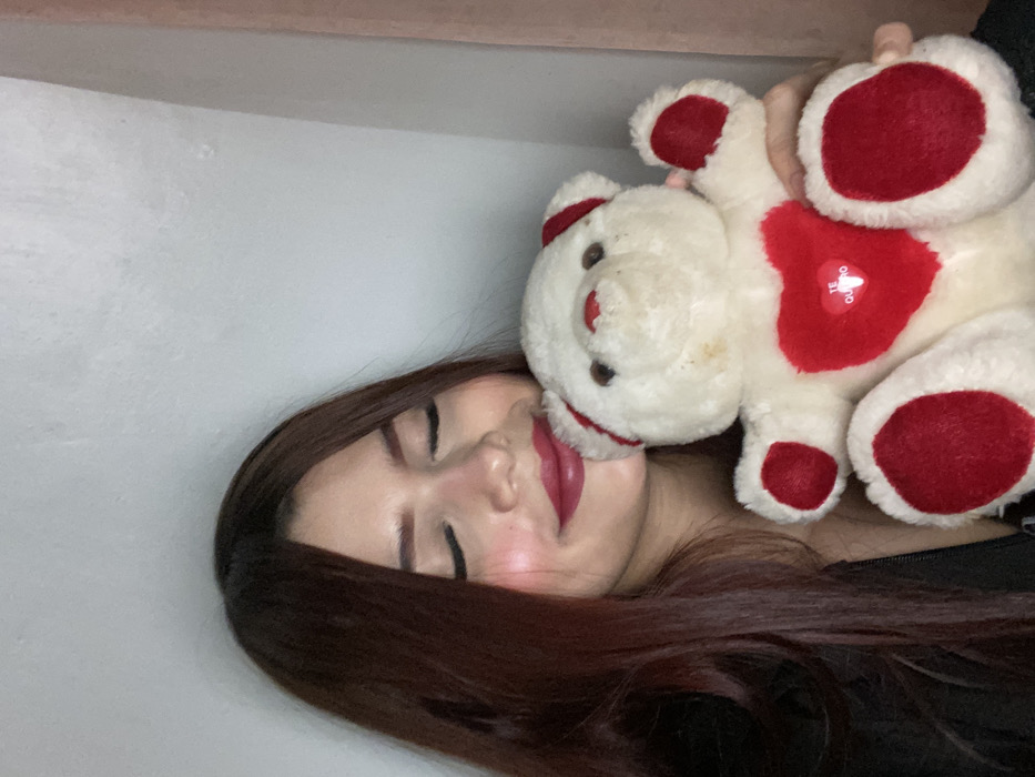 A woman with long, dark auburn hair is posing cheek-to-cheek with a plush teddy bear. The bear is cream-colored with red heart-shaped patches on its chest and paws, and it appears to be held by someone out of frame. The woman has fair skin, closed eyes, and wears dark lipstick. The background is a pale, solid color. The image is oriented on its side.