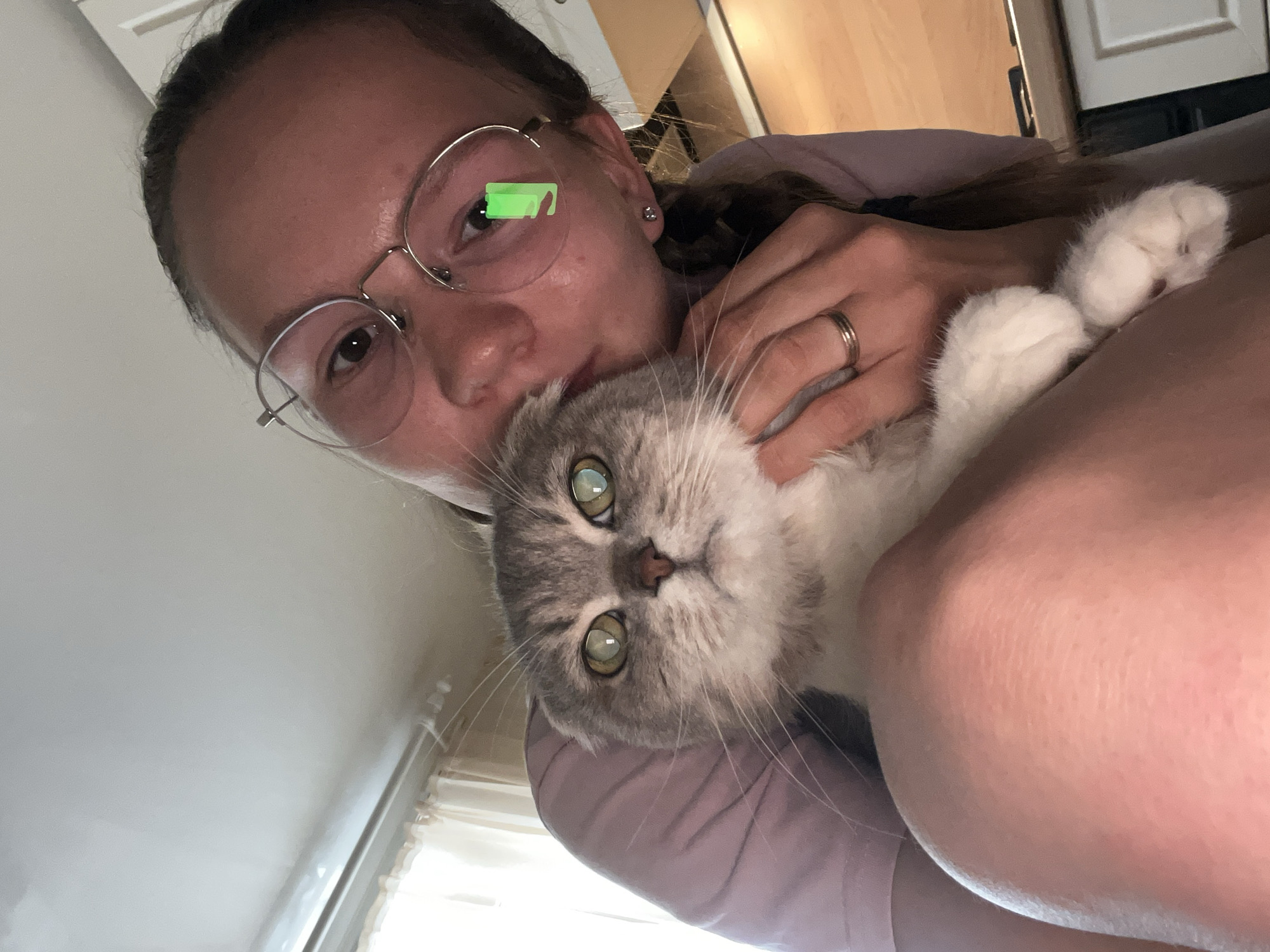 The image depicts a close-up, high-angle interior scene of a person holding a Scottish Fold cat. The individual is a female with light brown hair, possibly braided, wearing round, thin-rimmed metal eyeglasses. She has an earring in her right ear and a gold-colored band on her left ring finger. She is clad in a light purple top, with a portion of her bare knee visible in the lower left. She is positioned with her head leaned towards the cat's head, suggesting an act of kissing or nuzzling.
The cat, primarily grey and white with tabby markings on its face, has prominent green eyes that show a bright, yellowish-green reflection. Its distinctive folded ears are partially visible, confirming its breed. The cat's whiskers are long and its front paws are white and fluffy, visible at the bottom right of the frame. The cat appears to be looking directly at the viewer with a neutral expression.
A distinct green rectangular shape, resembling a smartphone battery icon, is reflected in the right lens of the person's eyeglasses, suggesting the photo was taken with a mobile device. The background elements indicate an indoor residential setting. A white ceiling is visible at the top, and a window with light-colored curtains is present on the left side, providing ambient light. On the right, hints of a light-colored wooden countertop and white cabinets or drawers are discernible, suggesting a kitchen or similar area. The overall lighting is bright and even.