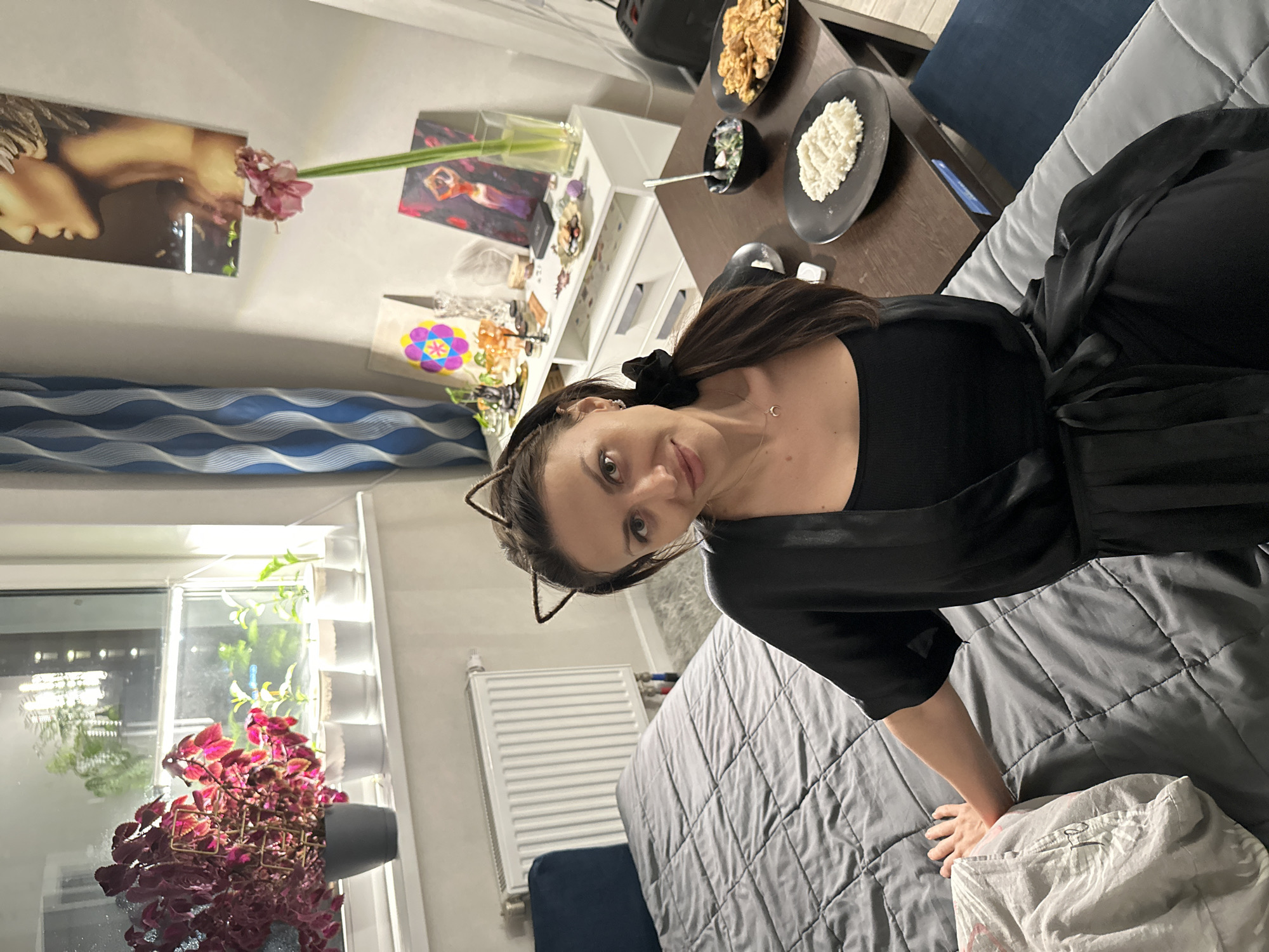 A woman is positioned near a bed in a room with various items. She has dark hair, fair skin, and is wearing a black top and a black satin robe with cat-ear headbands. The bed has a gray quilted bedspread. A small table is visible with plates of food and a bag. A window with plants and a radiator is to the left of the frame. Decorations, including a framed photograph, are visible on a shelf in the background. The location is identified as Murino, Russia.
