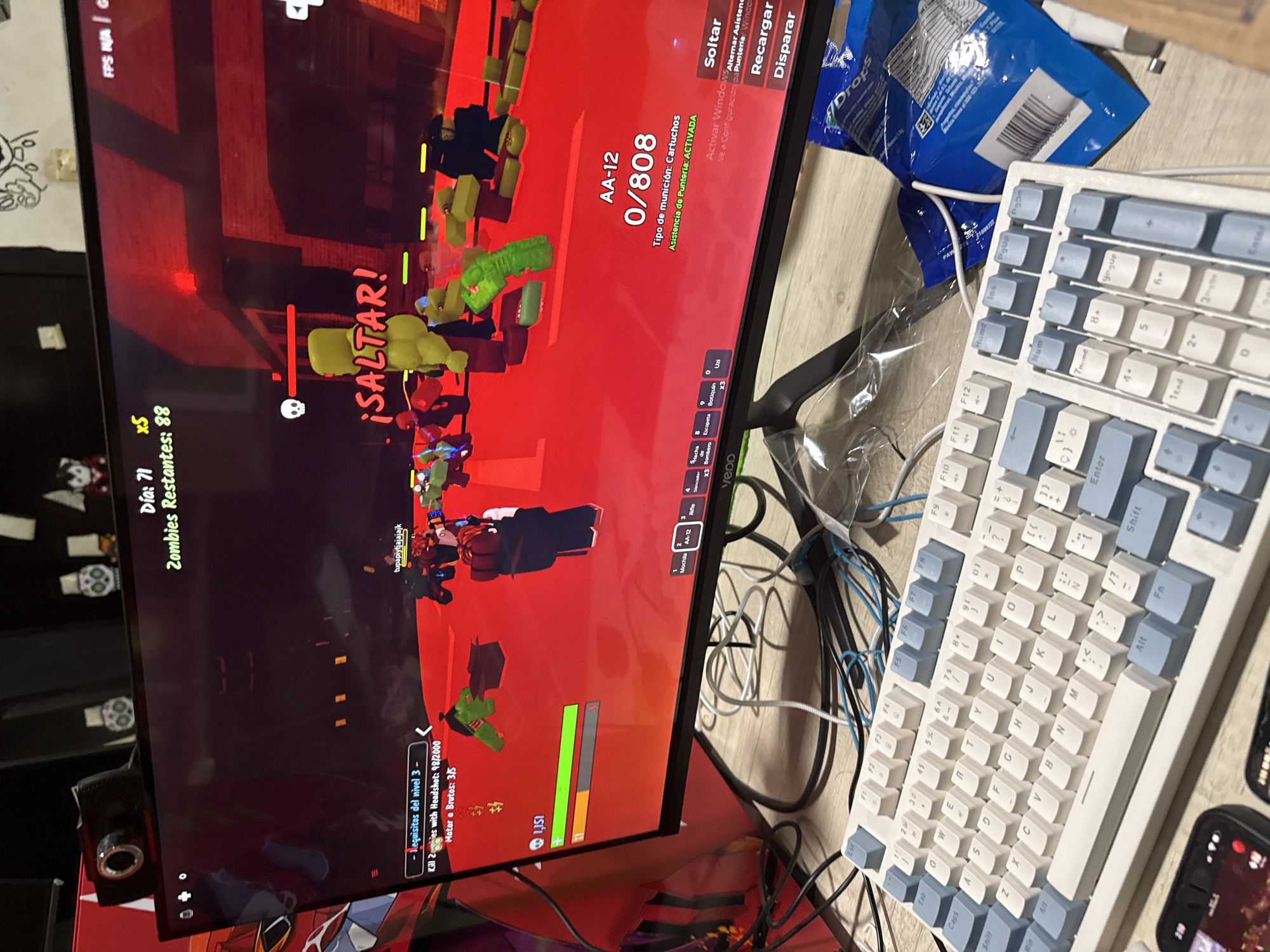 The image depicts a computer screen displaying a video game. The game appears to be a first-person shooter with zombies, indicated by the health bar, ammunition count (AA-12 0/808), and the text "Zombies Restantes: 88". The player character is in combat, as suggested by the "¡SALTAR!" (Jump!) prompt. The screen also displays game objectives: "Kill Zombies with Headshot: 98/2000" and "Matar a Brutos: 3/15".
To the right of the screen, there is a white and blue computer keyboard and a blue bag of chips labeled "Drops". Cables are tangled around the base of the monitor and keyboard. A smartphone is also visible on the desk.
The image does not contain information about the location of the city of Puebla, Mexico.