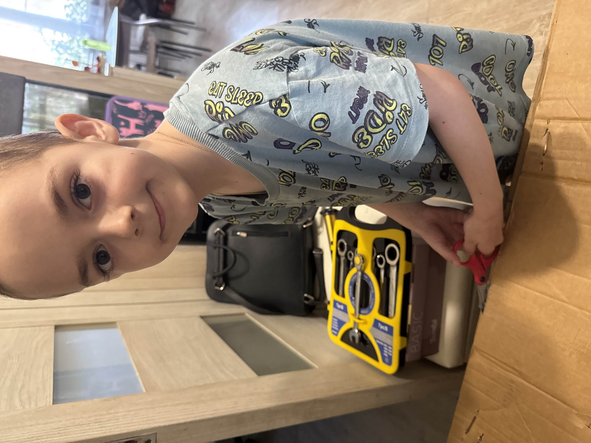 A child is cutting a piece of cardboard with red scissors. Next to them is a black backpack, a yellow tool set, and books. The location is unclear, but it could be in Раздолье, Russia.