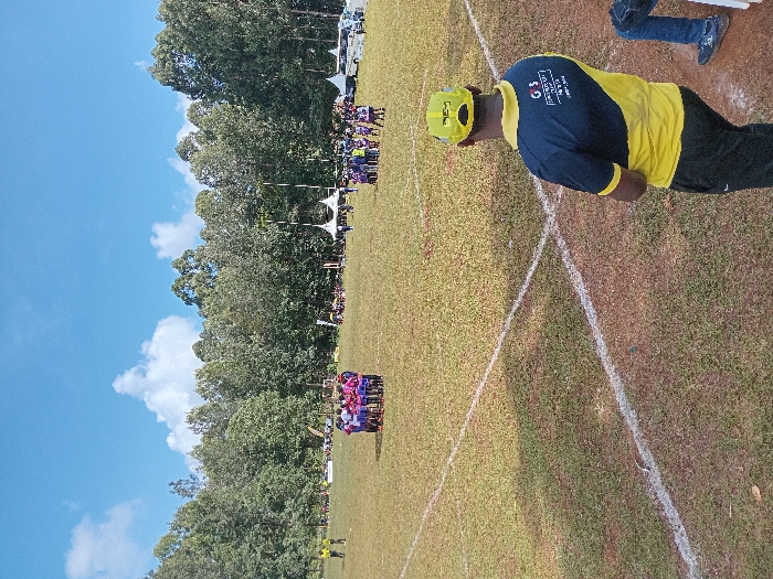The scene depicts an outdoor sports event on a grassy field in Meru, Kenya, under a blue sky with scattered cumulus clouds. In the lower right foreground, a male subject, viewed from the back, wears a yellow baseball cap and a two-tone navy blue and yellow polo shirt featuring "GS FINANCIAL TEP Meru" text. He stands near intersecting white lines painted on the grass. In the mid-ground, a group of approximately 10-15 individuals, primarily in pink/purple and dark sports uniforms, are gathered in a tight circular huddle on the left side of the field. To their right, a larger, more dispersed group of people in various sports attire congregates. Along the far perimeter of the field, several white canopy tents are erected, with a white bus parked among them. A dense line of tall trees forms the background, with two slender white structures, consistent with goalposts, visible above the tents.