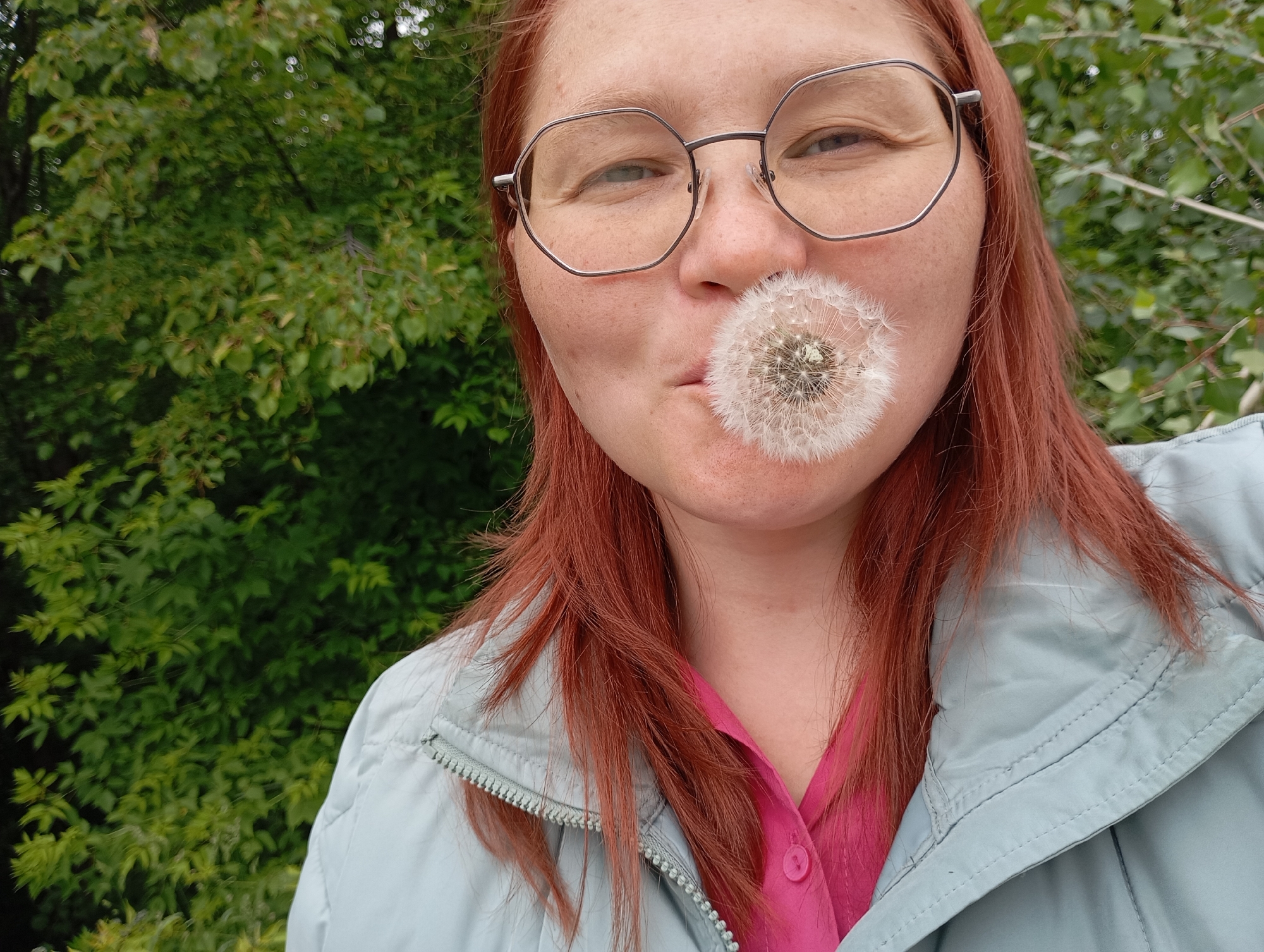 A close-up portrait shot shows a person with reddish-brown hair and glasses, holding a full dandelion seed head (puffball) to their mouth. The person is looking directly at the camera with a playful expression, eyes slightly squinted, and a hint of a smile. Freckles are visible across their nose and cheeks. They wear large, hexagonal-shaped metal-rimmed glasses.
The dandelion puffball is pristine, with all its fluffy white seeds intact, positioned as if the person is about to blow on it. The person's attire includes a light blue or pale grey zippered jacket worn over a bright pink collared shirt.
The background is a soft blur of dense green foliage, suggesting an outdoor, natural setting like a park or garden, consistent with the location of Chaikovsky, Russia. The lighting is soft and diffused, indicating an overcast day or a shaded area. No text is visible in the image.
