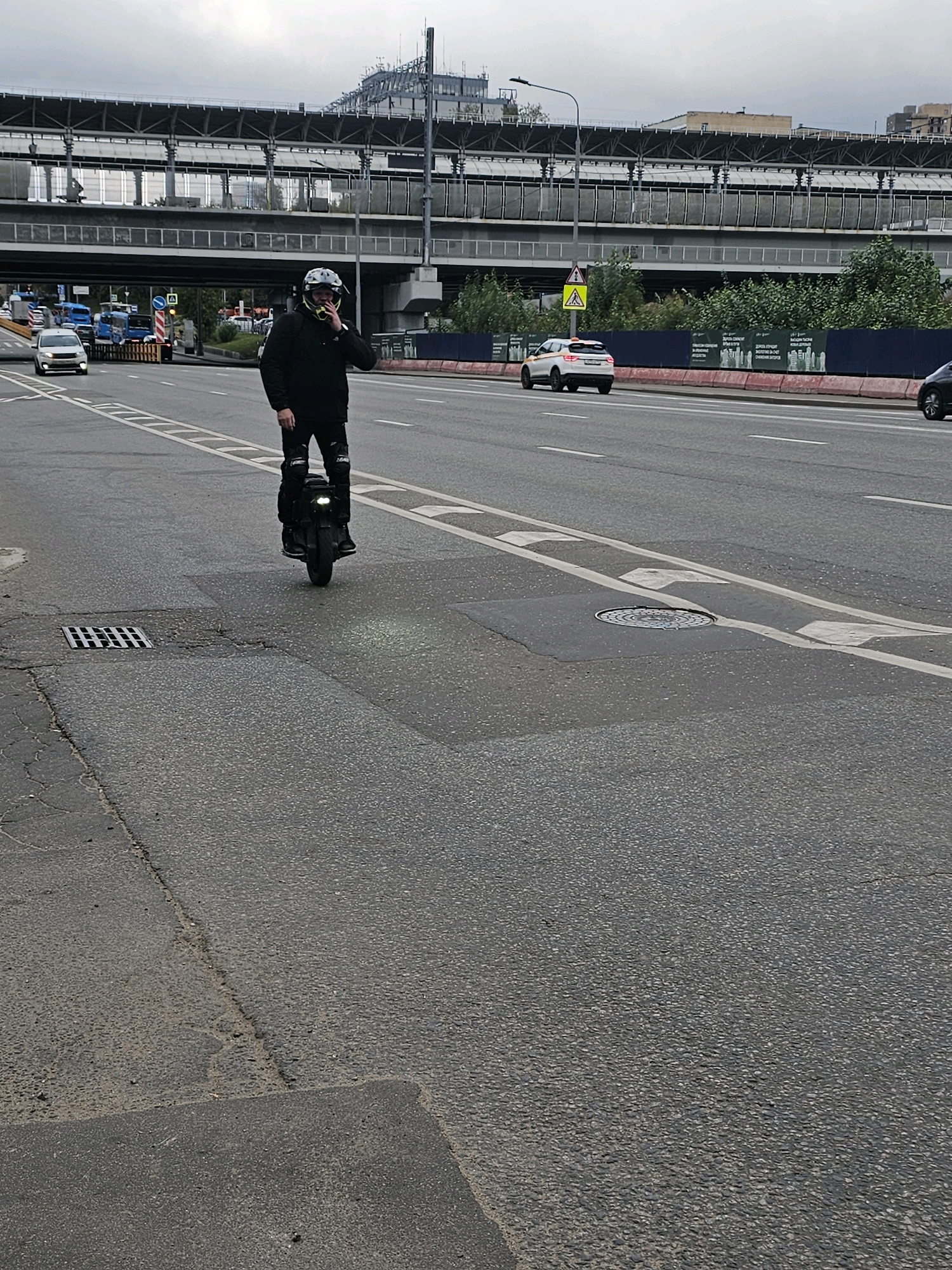 A person wearing a helmet and protective gear is riding a single-wheeled electric unicycle on a multi-lane road in Moscow, Russia. The person is holding a mobile phone to their ear and appears to be speaking. They are dressed in dark clothing, including knee pads. The unicycle has a headlight and a taillight.
The road is paved with asphalt and marked with white lane lines and chevrons indicating a traffic separation zone. There are cars and buses on the road in the background. Above the road, there is an elevated structure, likely a railway overpass or a highway, with visible support beams and a roof. Trees and buildings can be seen beyond the overpass. The sky is overcast. The scene appears to be in an urban environment.