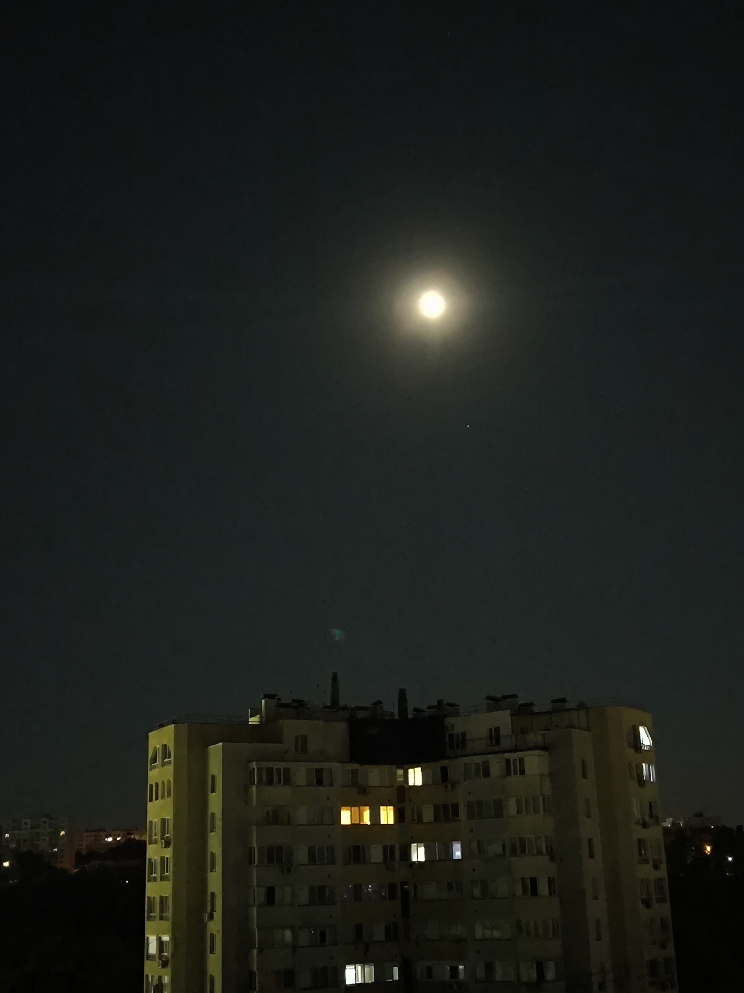 The image captures a nighttime scene in Chișinău, Moldova, dominated by a large, illuminated apartment building. A bright full moon hovers in the dark sky above, casting a soft glow. The apartment building, likely a Soviet-era residential block, features numerous windows, many of which are lit from within, indicating occupancy. Some windows appear to be balconies. The building is bordered by dark trees and other distant buildings with faint lights. The overall impression is of a quiet urban evening, with the moon serving as a prominent celestial body.