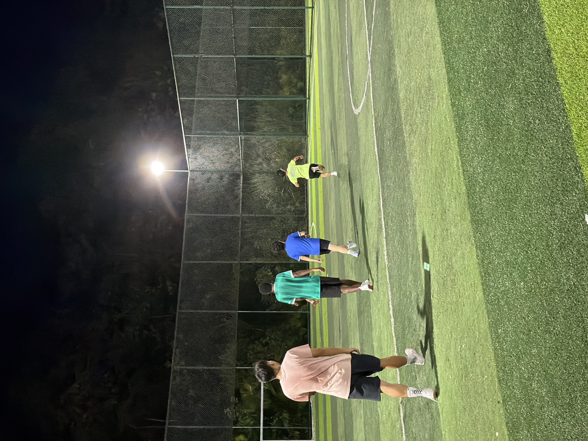 Four young males are on a brightly lit, outdoor artificial turf soccer field at night. A tall chain-link fence encloses the field, and lush vegetation is visible behind it. The sky is dark, and a single, powerful floodlight illuminates the scene from above. The individuals, viewed from behind, are engaged in activity on the field. One individual in a pink t-shirt and dark shorts is positioned on the left side of the frame, facing away from the camera and towards the center of the field. To his right, another individual in a green t-shirt and black shorts walks away from the camera. Further to the right, two more individuals are running across the field. One is in a blue t-shirt and black shorts, while the other wears a neon yellow t-shirt and black shorts. The field has visible white markings for boundaries and circles. The overall impression is of an active evening sports gathering in an urban or suburban park setting within Jiangjin, China, under the illumination of artificial lights.