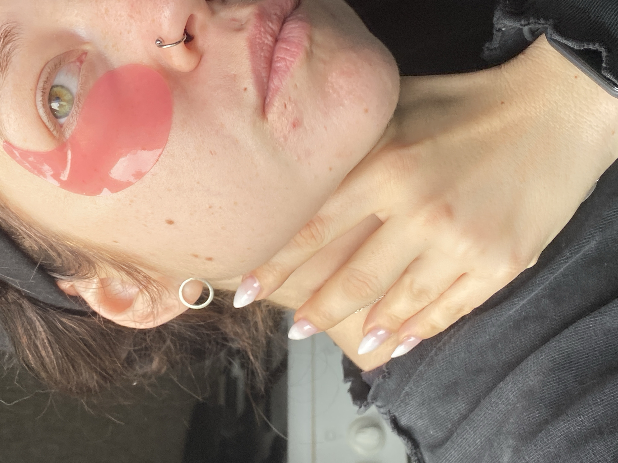 A close-up, eye-level forensic photograph depicts a light-skinned individual, likely female, from the mid-chest upwards, focusing on the right side of their face and neck. The individual has brown hair, partially obscured by a dark headband, visible on the right side of the frame. A single green eye is prominent, with a pink, teardrop-shaped hydrogel eye patch positioned directly beneath it. The skin exhibits scattered freckles and minor blemishes, particularly around the chin and lower lip area. The visible left earlobe features a simple, silver-colored hoop earring. A silver-colored twisted hoop nose ring is present in the left nostril. The individual's left hand, with almond-shaped fingernails featuring a white-to-clear ombre French manicure, is gently resting against the right side of their neck, just below the jawline. The visible attire is a dark, possibly charcoal or black, ribbed or textured garment, with a somewhat distressed neckline. The background is indistinct and out of focus, primarily dark, with some blurry lighter elements suggesting an indoor environment. There is no discernible information within the image to indicate the city of Chaikovsky, Russia, or any specific geographical location.