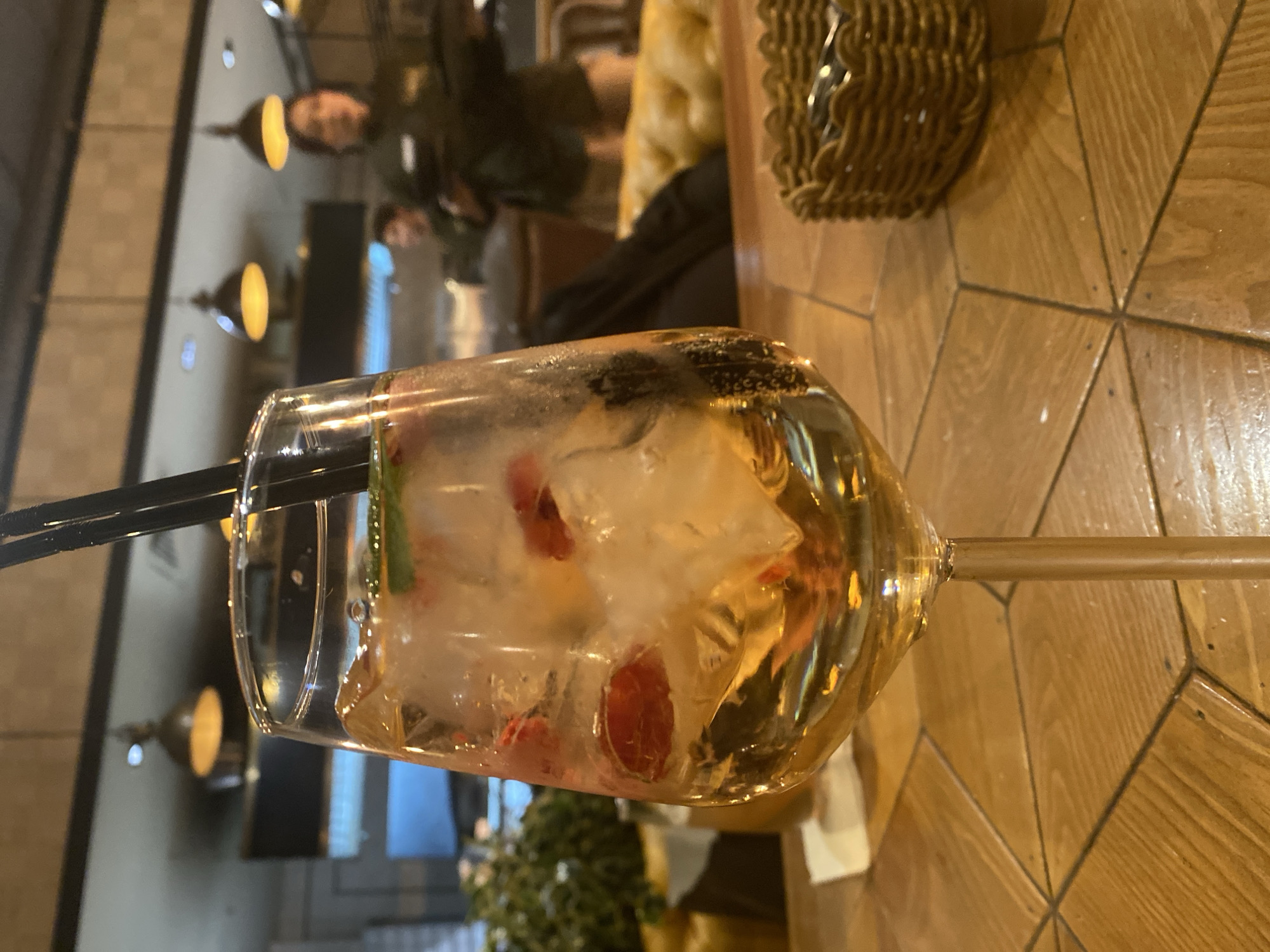 A close-up, high-angle shot shows a stemmed glass filled with liquid, ice, and fruit. Two black straws are inserted into the glass. The drink is placed on a wooden table with a geometric pattern. In the background, blurred figures of people are visible. The background also shows hanging light fixtures. The location could be a cafe or restaurant in Yekaterinburg, Russia, based on the presence of Cyrillic script in the background.
