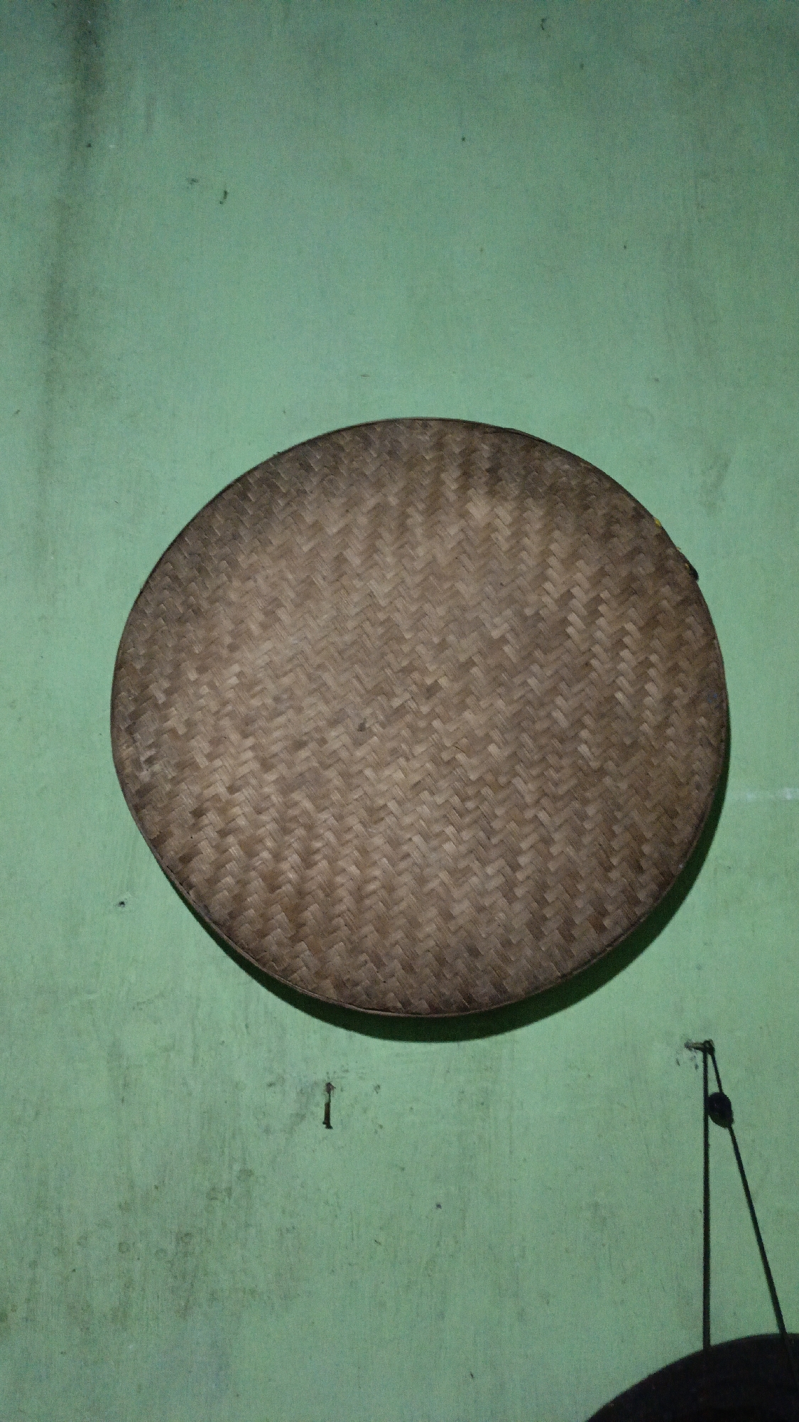 The image displays a woven, circular object, likely a tray or a sieve, hanging against a light green, textured wall. The object is made of a natural material, possibly bamboo or rattan, with a herringbone weave pattern. Its color is a warm brown, with variations in shade due to the natural material and lighting. The wall exhibits minor imperfections, including some faint dark smudges and a small, dark object resembling a nail or hook directly below the woven item. To the right of the woven object, another dark object, possibly part of a hanging mechanism or another decorative item, is partially visible. The lighting in the scene appears to be from an artificial source, casting a subtle shadow around the woven object and on the wall. The overall impression is one of a rustic, domestic setting.
