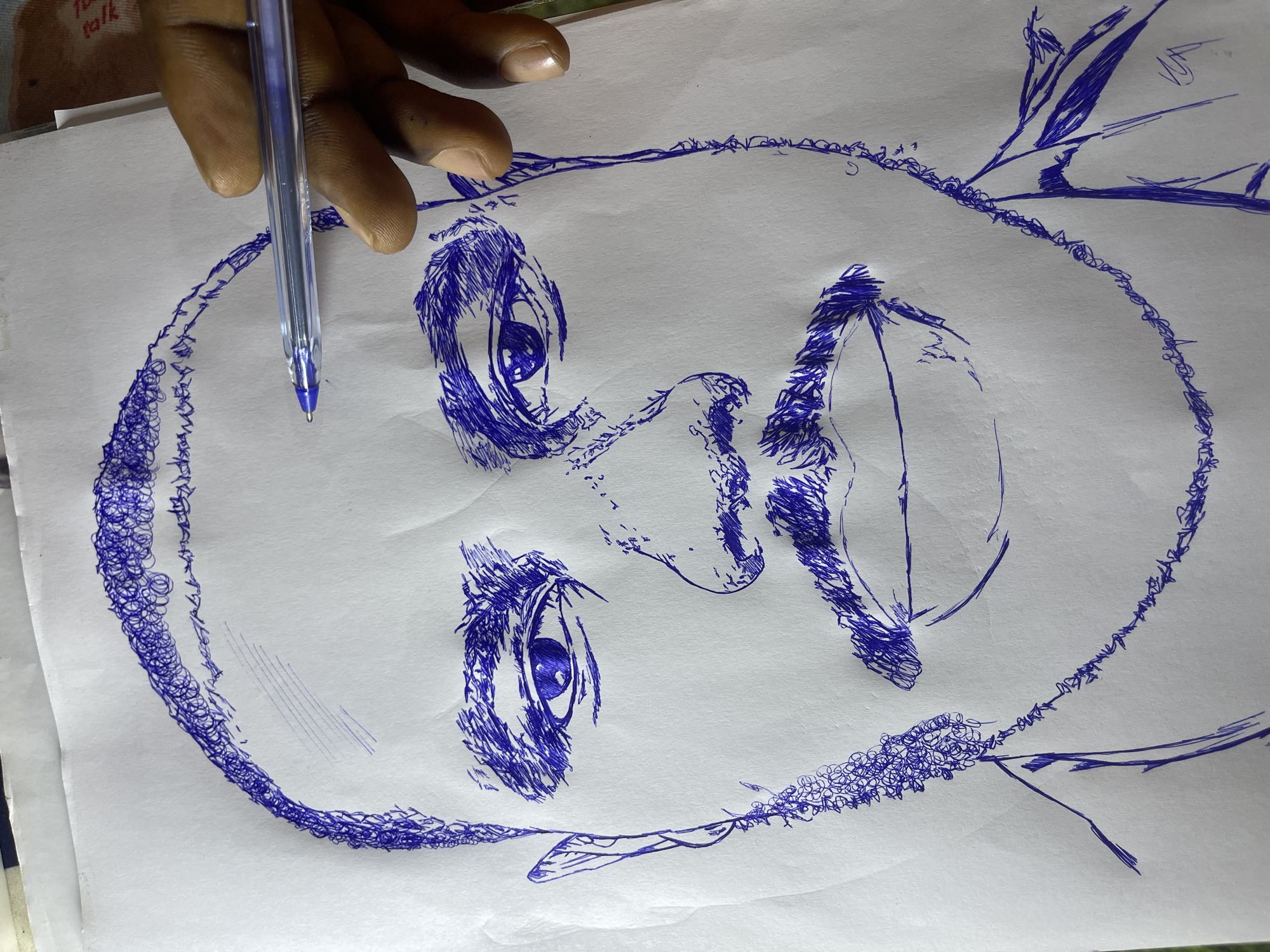 The image displays a pen drawing of a man's face on a white sheet of paper. The drawing is rendered in blue ink using a cross-hatching and scribbling technique. The man has a bald head, prominent eyebrows, a mustache, and a goatee. His eyes are open, and he has a neutral expression. A portion of a hand holding a blue ballpoint pen is visible in the upper right corner of the image, suggesting the drawing is in progress. The background is plain white paper. The image is not depicting a scene in the city of Jos, Nigeria; it is a close-up of a drawing.