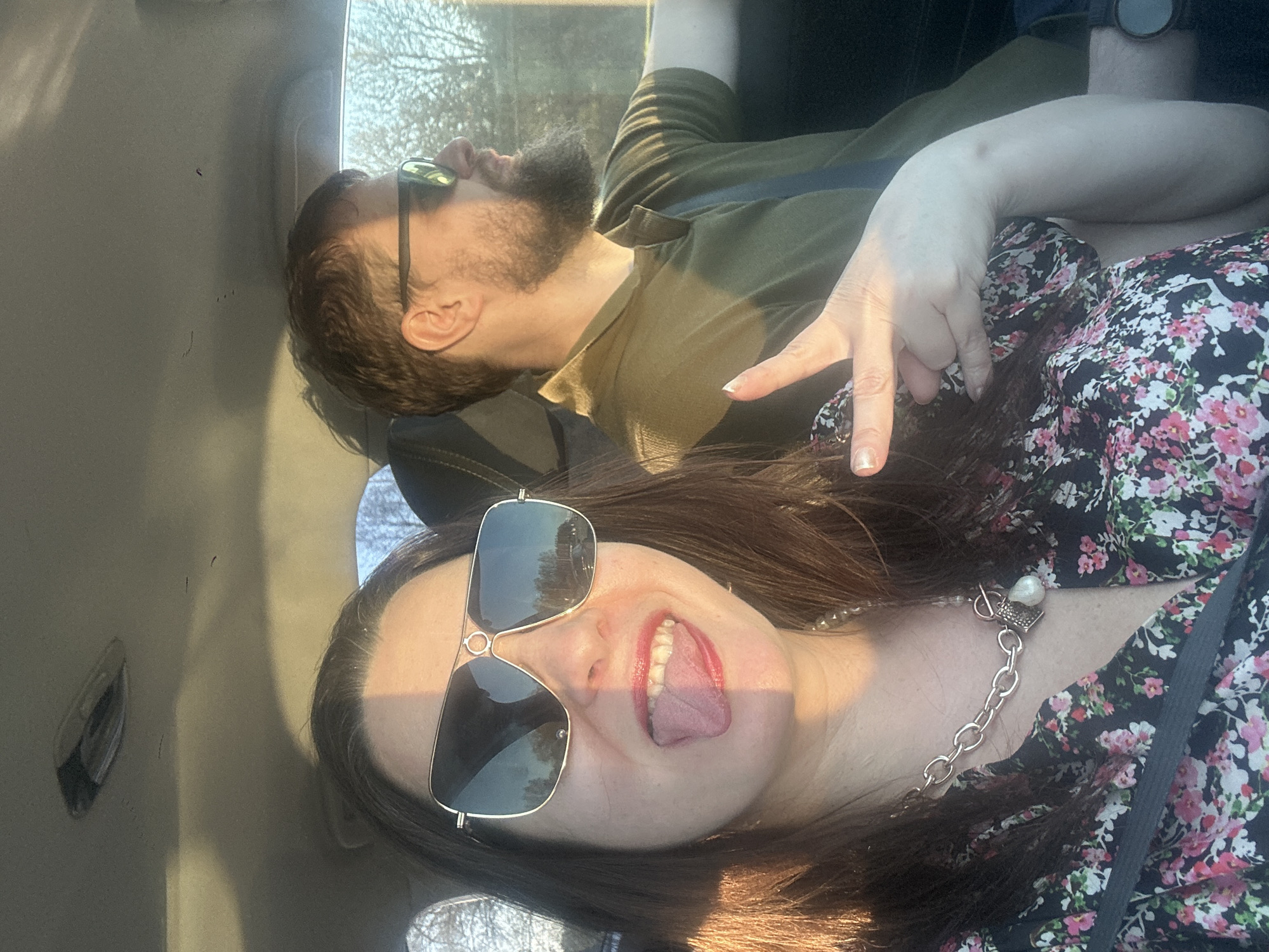 Two people are in a car. A woman in the foreground is wearing large sunglasses, a floral shirt, and a silver chain necklace. She is sticking her tongue out. A man behind her is wearing sunglasses and a green shirt. A hand is raised in a peace sign. The location is unknown.