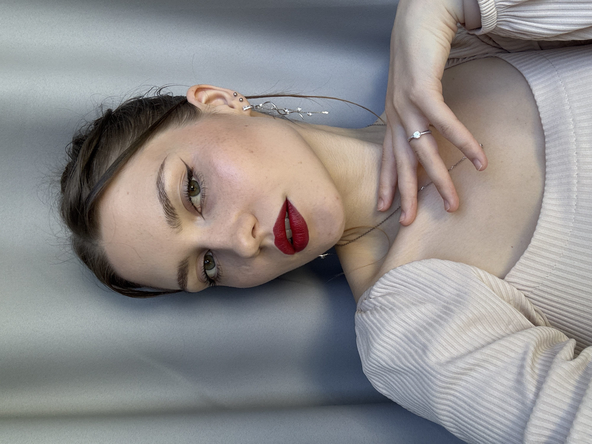 A young adult female is depicted in a close-up portrait, looking directly at the viewer with a neutral expression. Her dark brown hair is styled in an updo with some wisps framing her face and a longer strand falling over her right shoulder. She has green eyes, defined dark eyebrows, and wears prominent red lipstick with a matte finish. Black winged eyeliner is applied to both eyes, and her complexion is fair with visible freckles or small moles on her cheeks.
She is wearing a light beige or cream-colored top with a distinct ribbed texture and puffed sleeves, featuring a wide, possibly square or boat, neckline. Her left hand is gently placed on her upper chest/collarbone area. On her left ring finger, she wears a silver-toned ring with a single clear circular stone set between smaller stones. A thin silver-toned chain necklace with a small pendant is visible around her neck, partially obscured by her hand.
Her right ear displays multiple silver-toned earrings: two small studs and a long, ornate dangle earring featuring a twisted design adorned with multiple small, clear gemstones. A single stud earring is visible on her left ear.
The background consists of a plain light gray or blue-gray fabric, exhibiting a subtle vertical woven texture, strongly suggesting a curtain or a photographic backdrop. The lighting appears soft and even, illuminating the subject from the front. The image does not contain any visual information to determine a specific geographical location, such as Saint Petersburg, Russia.